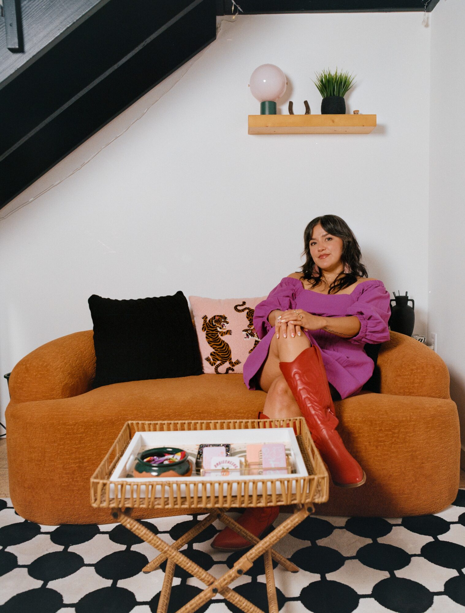 Woman sitting on a brown sofa with black and patterned pillows, in a room with black and white patterned floor and white wall, shelf with decor above.