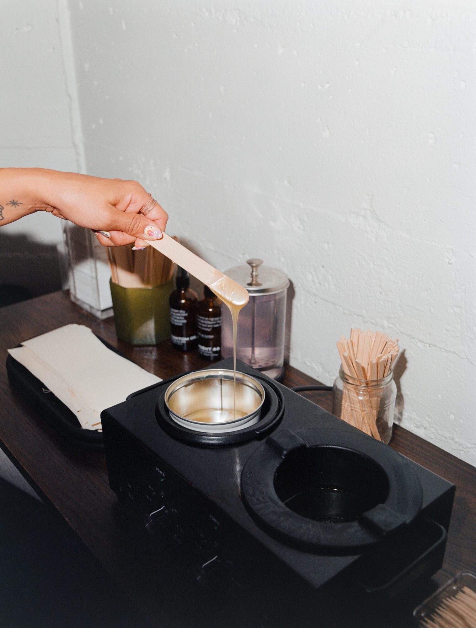 Hand holding a wooden spatula over a container on a black countertop with various bottles and a container of toothpicks, against a light-colored wall.