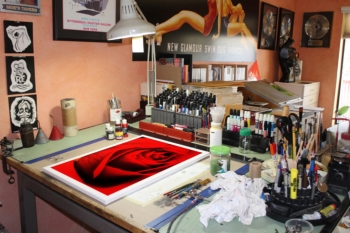 Workstation with art supplies, a digital tablet displaying a red rose, and various tools on a desk against a peach wall.