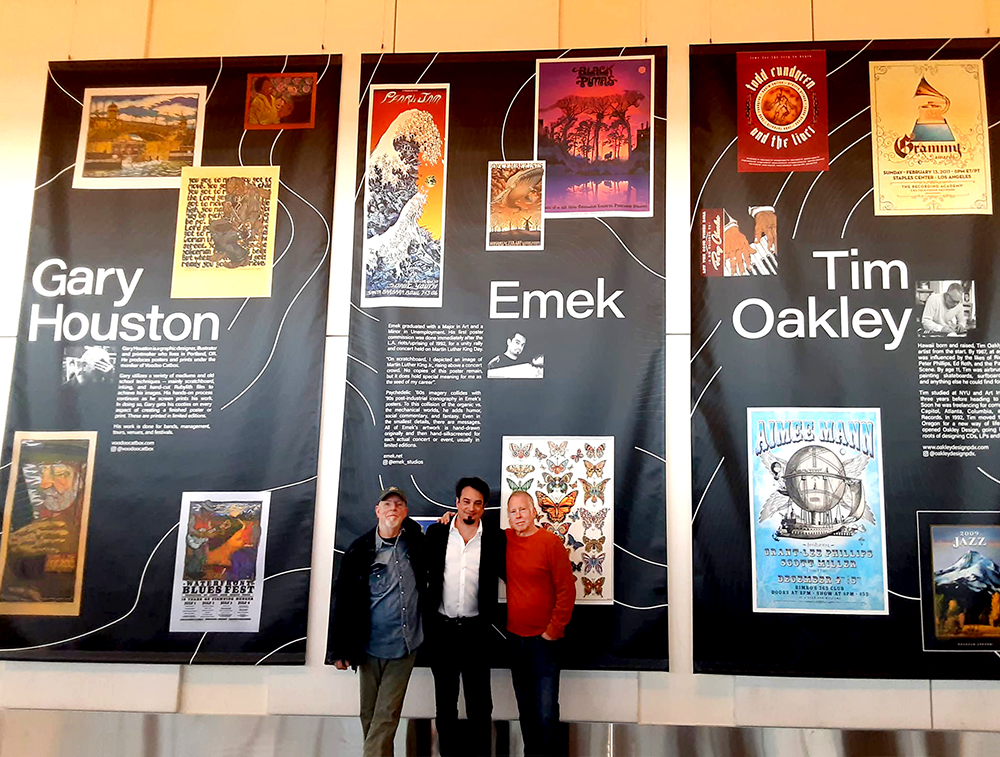 Three people stand in front of large display panels with artwork and text, indoors. The panels are black with colorful images.