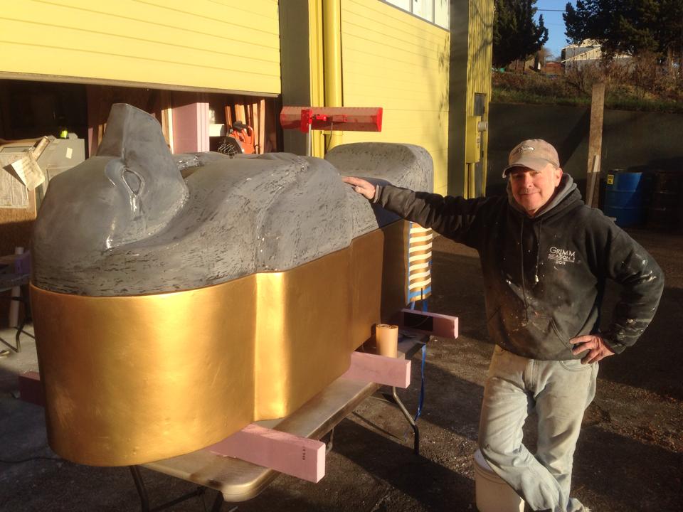 Person in a black jacket and beige cap standing next to a large gray sculpture of a sleeping animal with a gold base, outdoors.