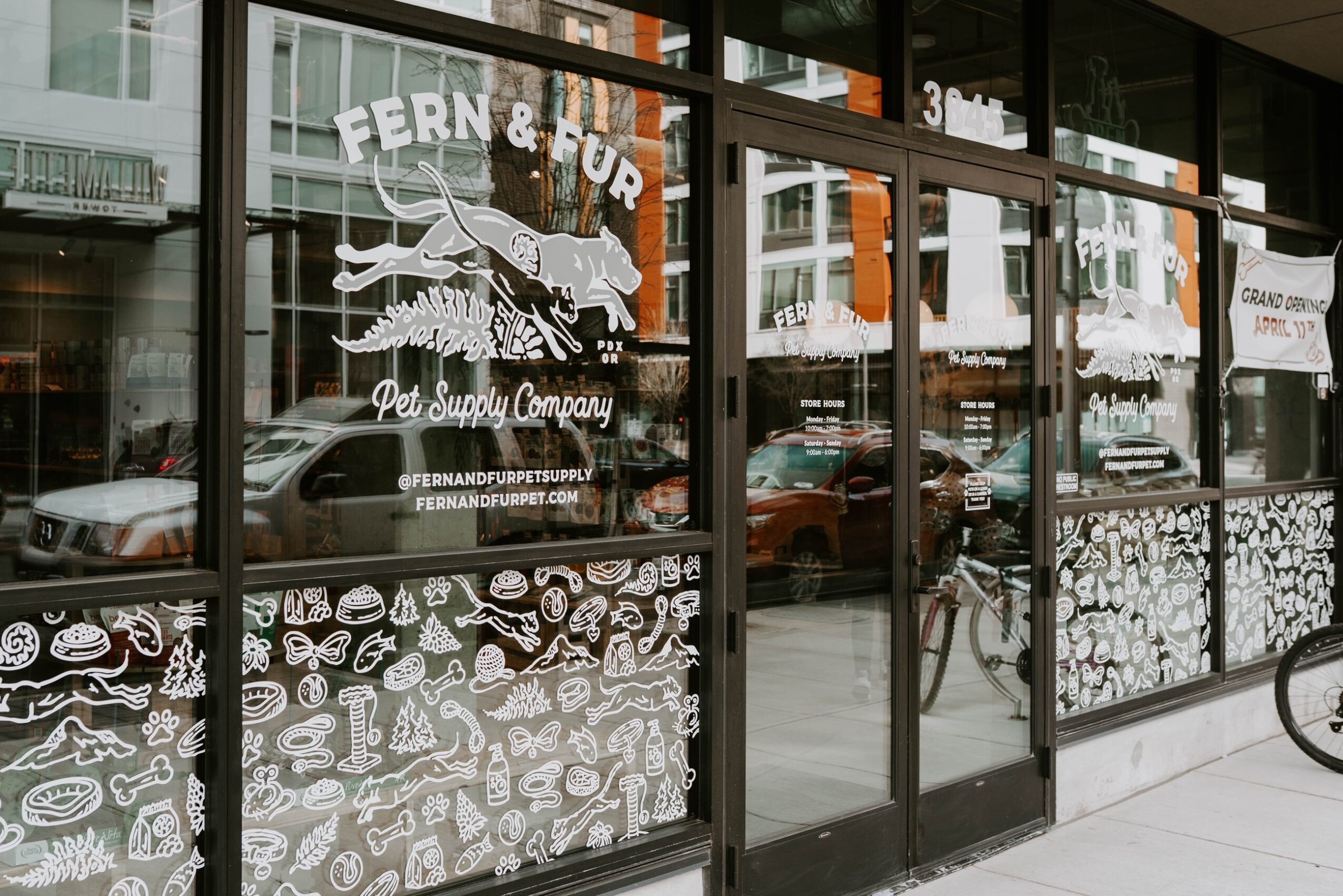 Storefront with large glass windows displaying pet supply company logo and illustrations, street view with parked cars and bicycles.