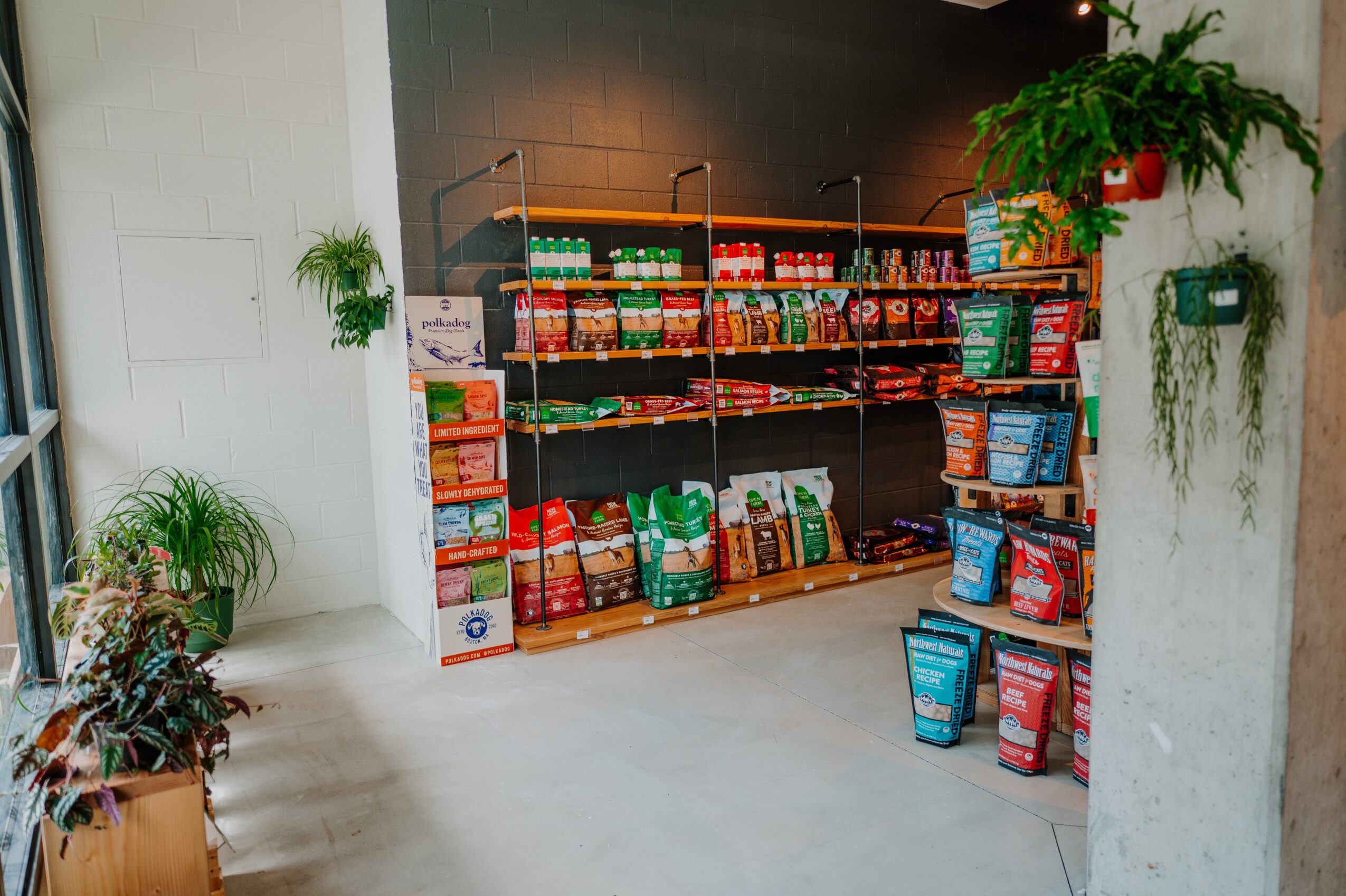 Shelves stocked with various food products in a store, surrounded by potted plants on white walls and floor.