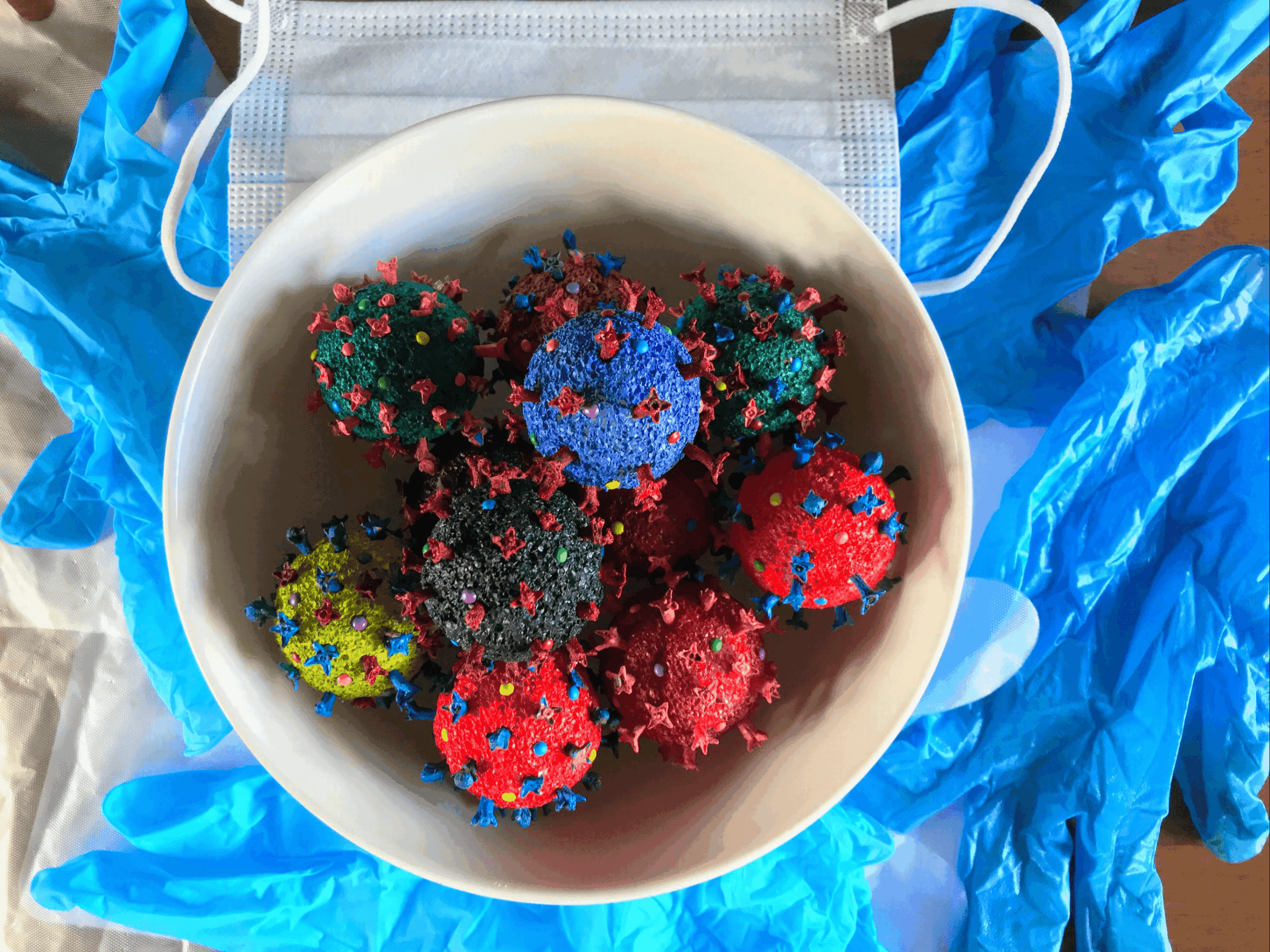 A white bowl contains colorful molecular models with red, blue, green, yellow, and black components, surrounded by blue medical gloves and a face mask.