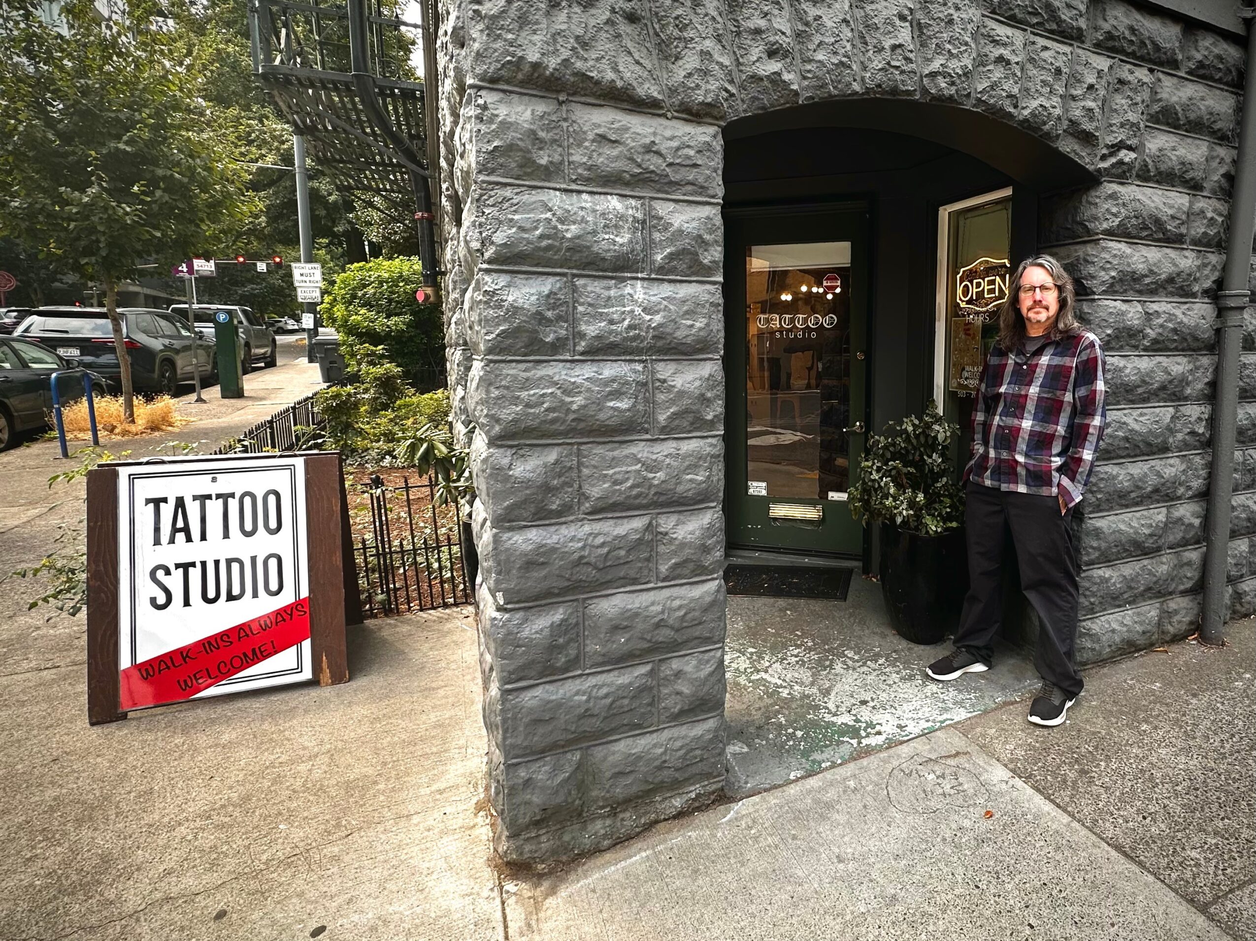 Person standing outside tattoo studio entrance with a sign on sidewalk, stone building facade, and greenery nearby.