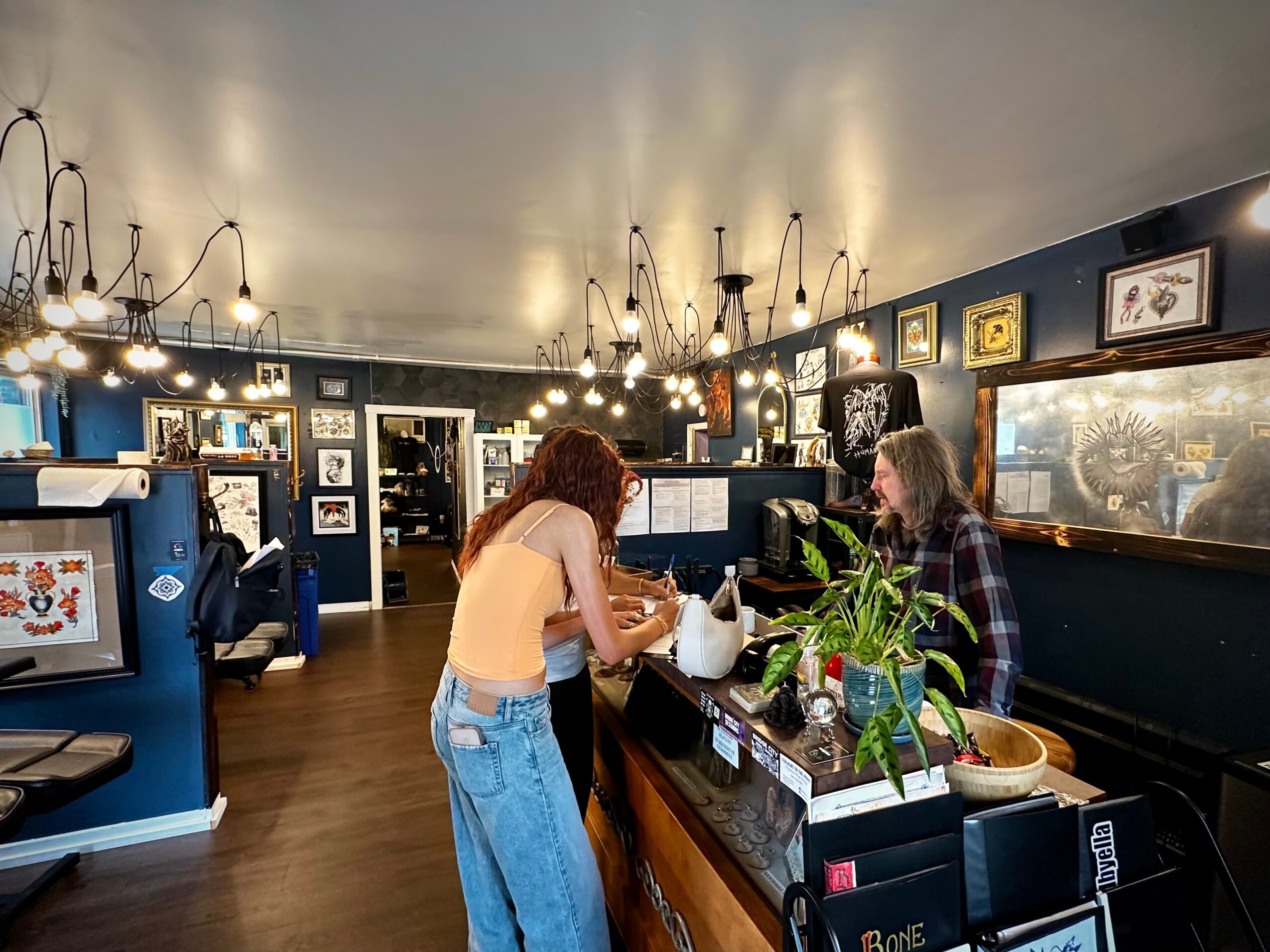 Two women at a bar counter, one with curly hair and the other with straight hair, engaging in conversation. The bar has framed pictures and plants.