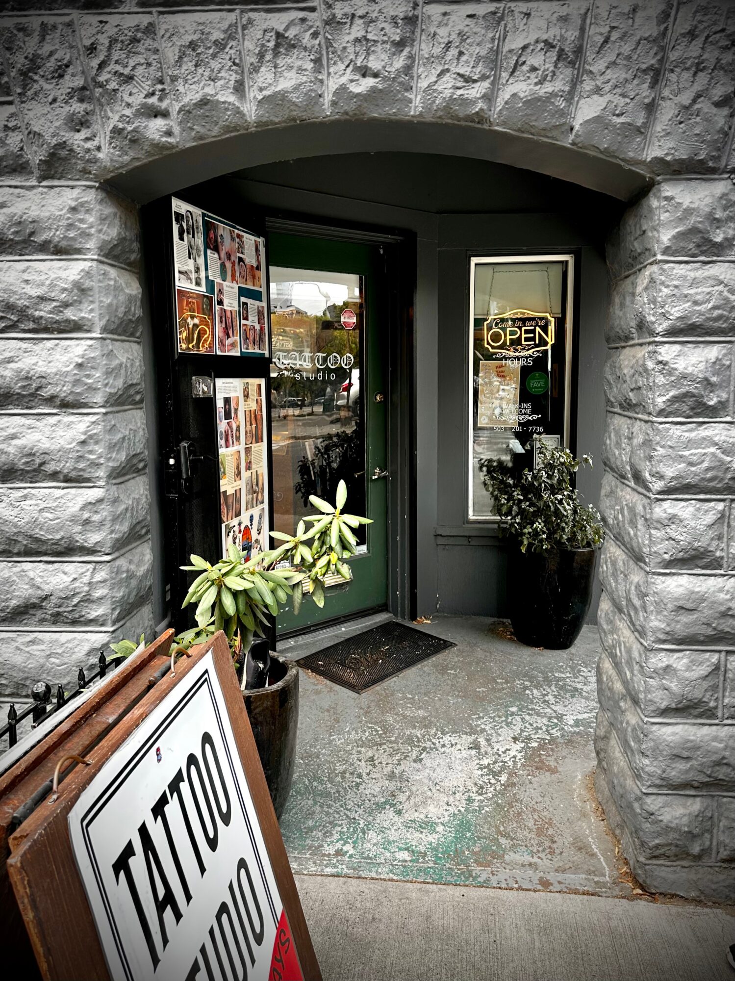 Stone archway entrance with a door, window, potted plant, and a sign for a tattoo studio outside.