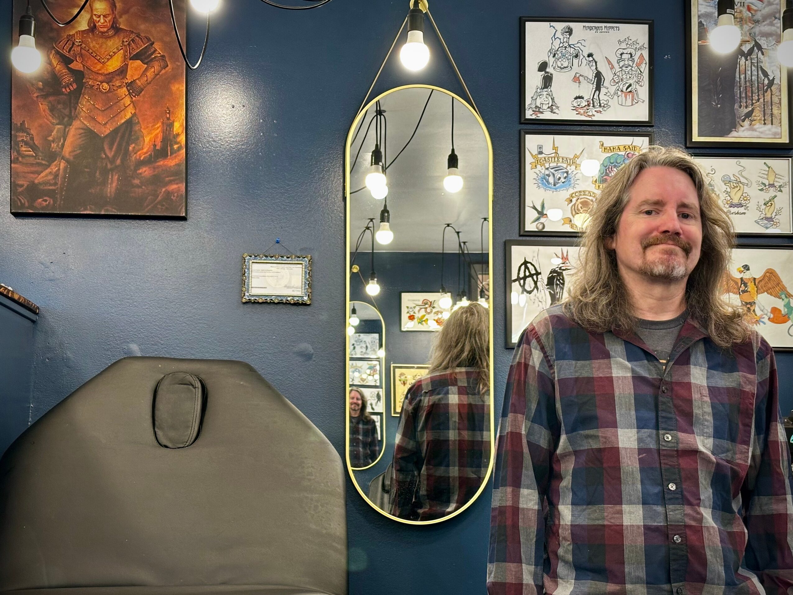 Man with long hair and beard wearing a plaid shirt, smiling, standing in a room with artwork and a mirror.