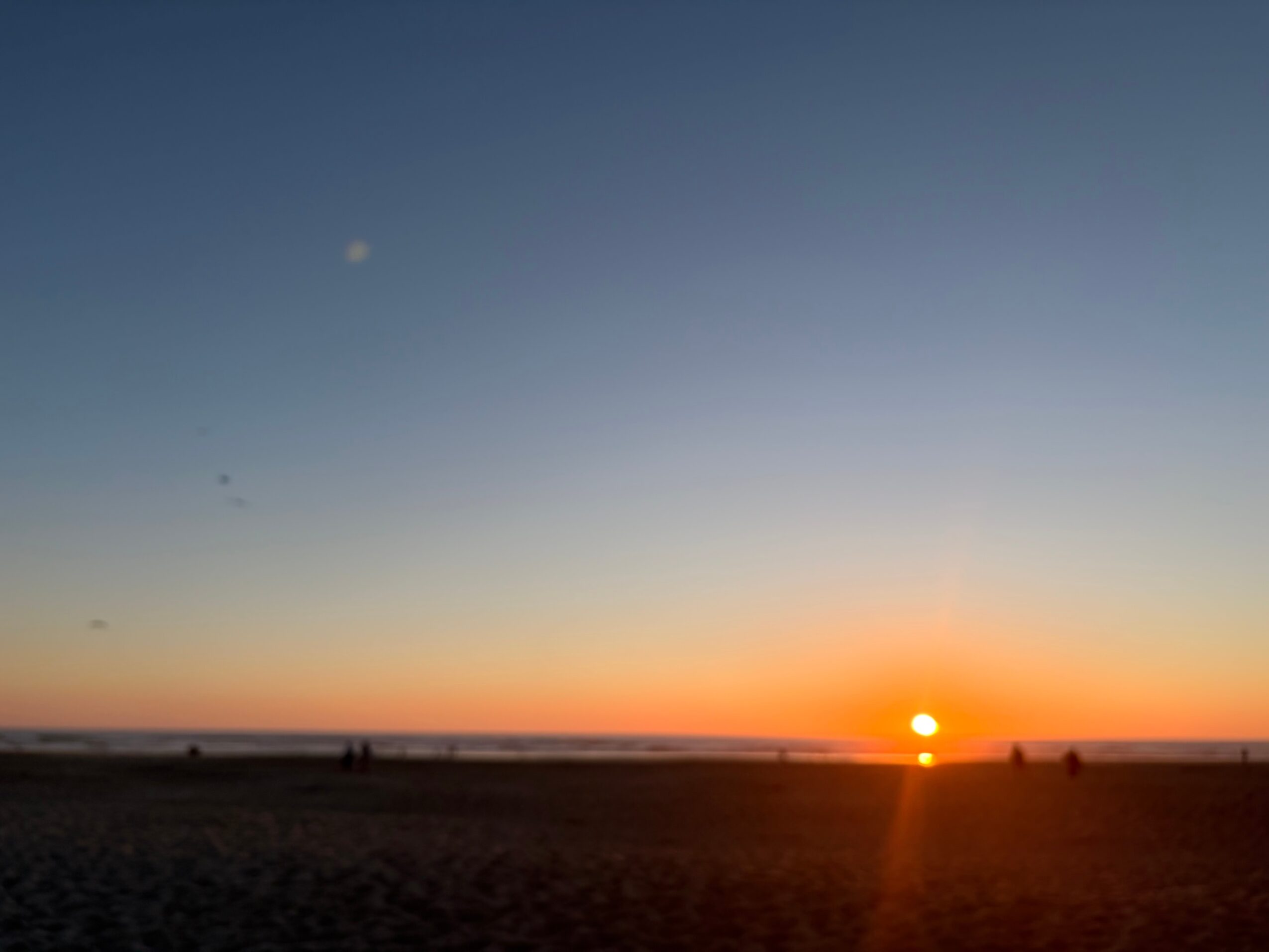 Sunset over the ocean with a clear sky, visible moon, and silhouettes of people on the beach.