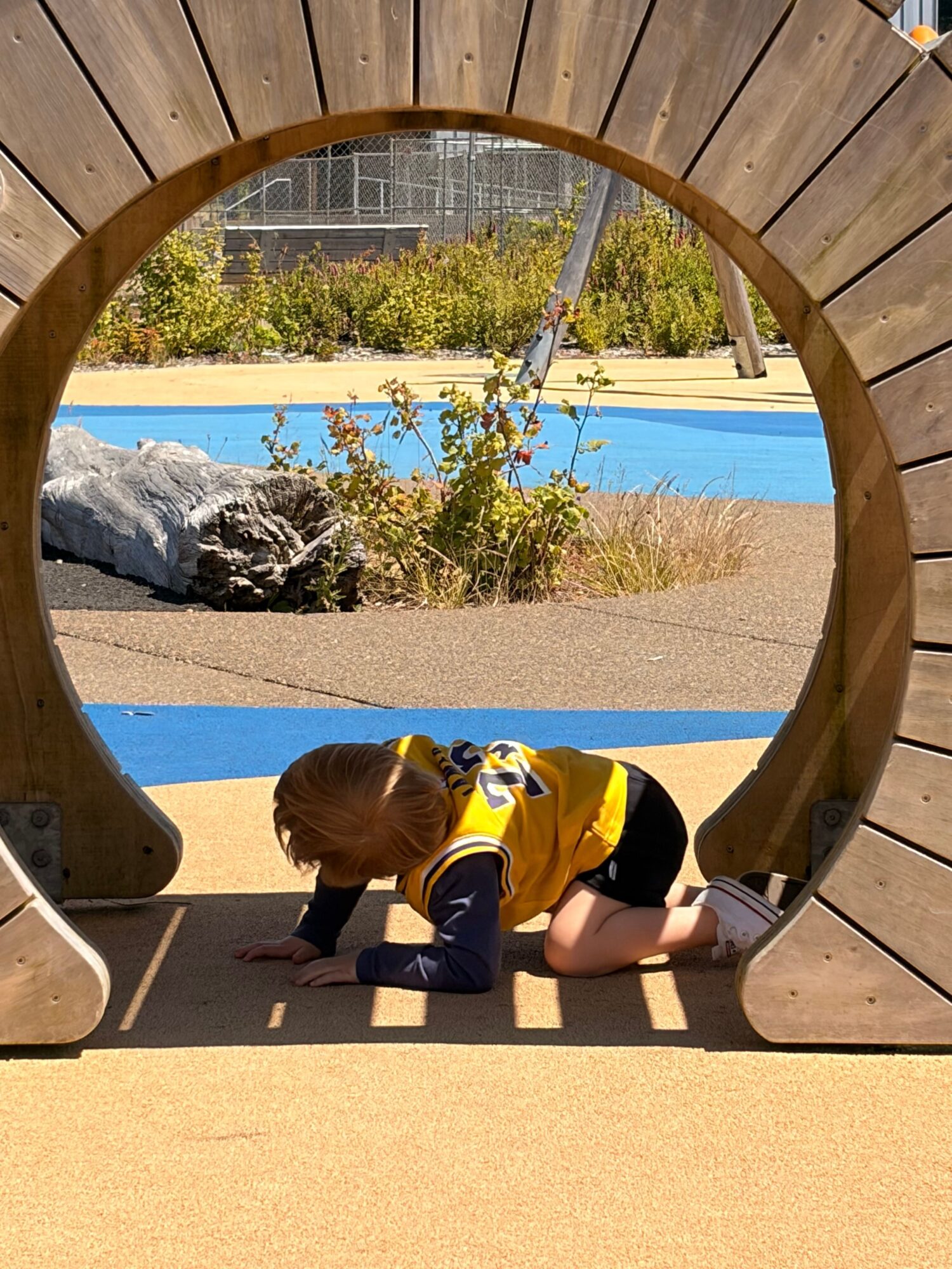 Child playing on a yellow and black toy car under a wooden circular structure, with a blue pathway and rocks in the background.