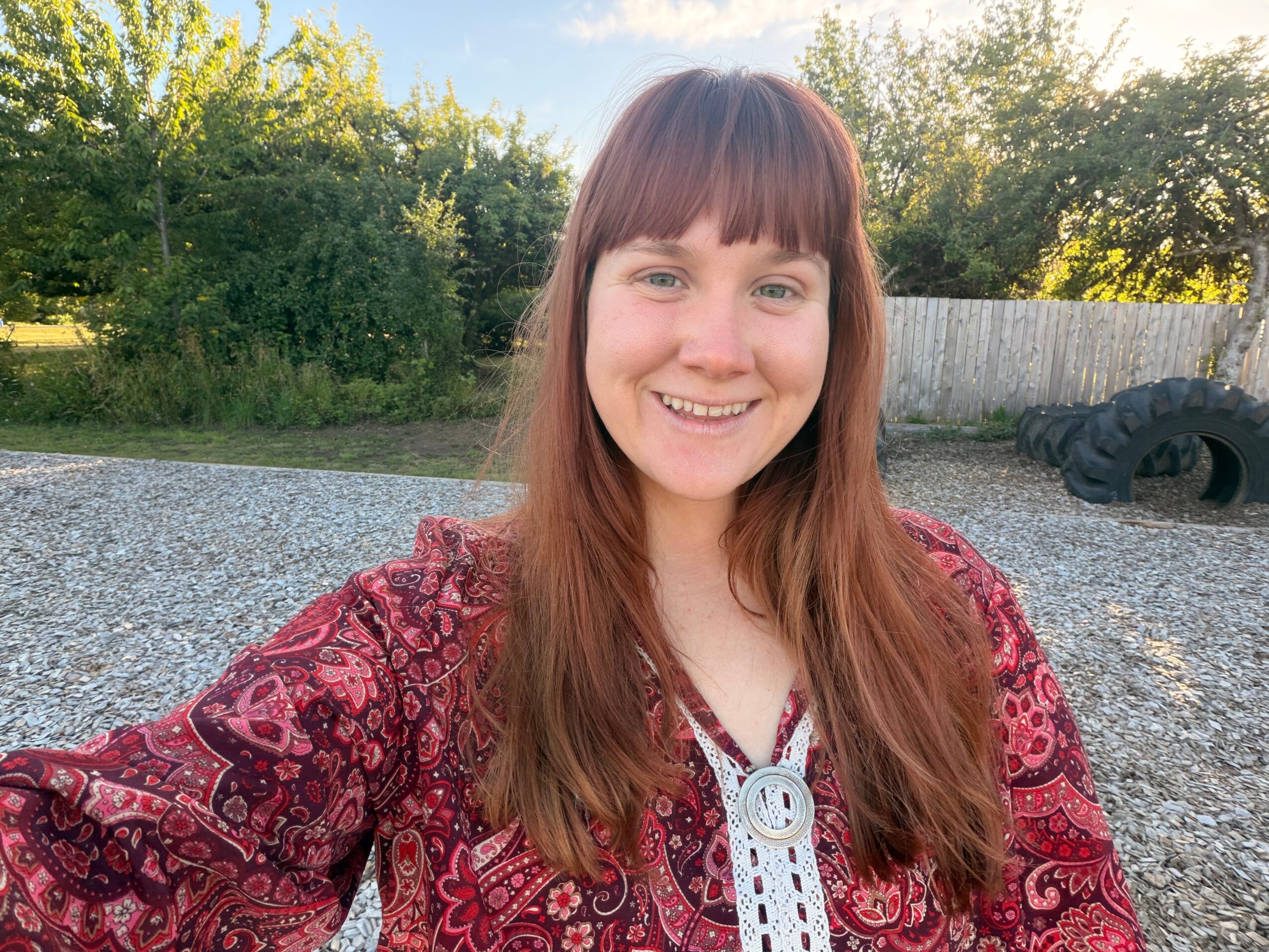 Smiling woman with long brown hair outdoors, wearing a red patterned jacket and white top, with trees and a tire in the background.