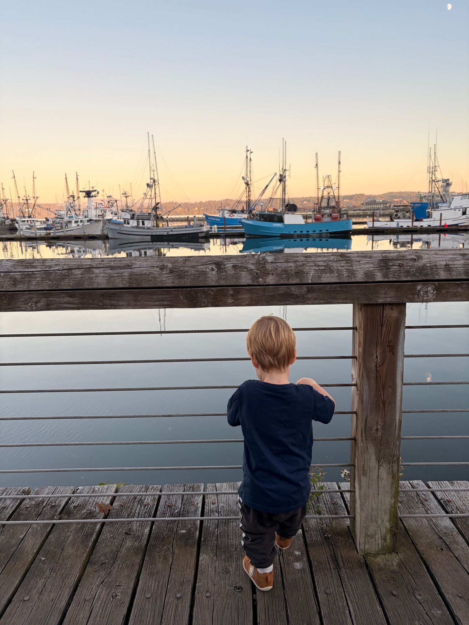 Child with blonde hair wearing a dark jacket and pants stands on a wooden dock looking at boats in the water, sunset sky.