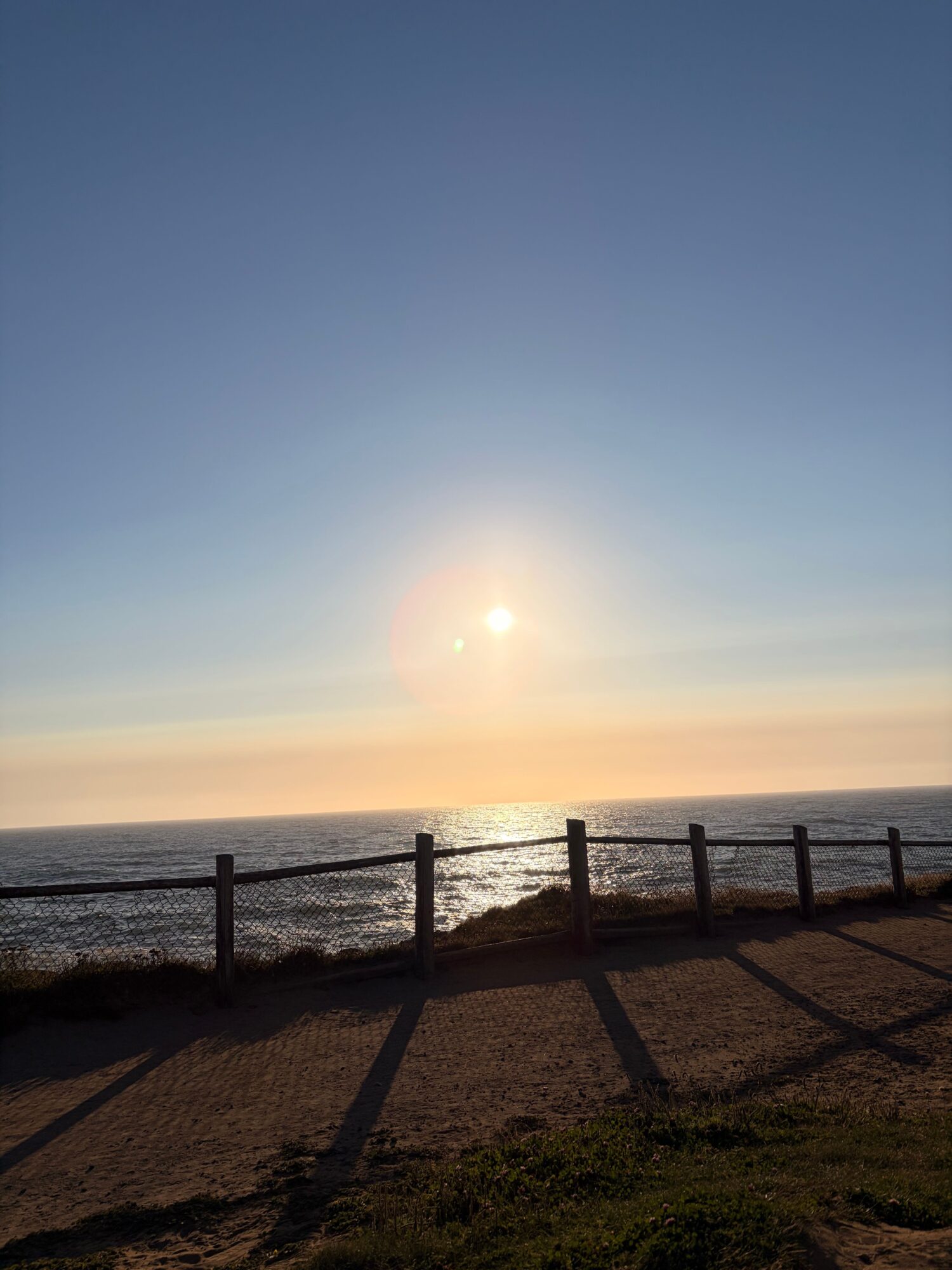 Sunset over the ocean with a wooden fence on the beach, shadows cast on sand, sky transitioning from blue to orange.