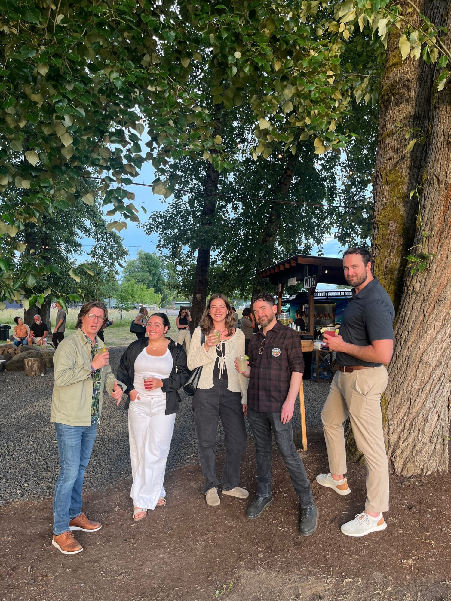 Group of six people standing outdoors near trees, posing for a photo, some smiling, in casual clothing.