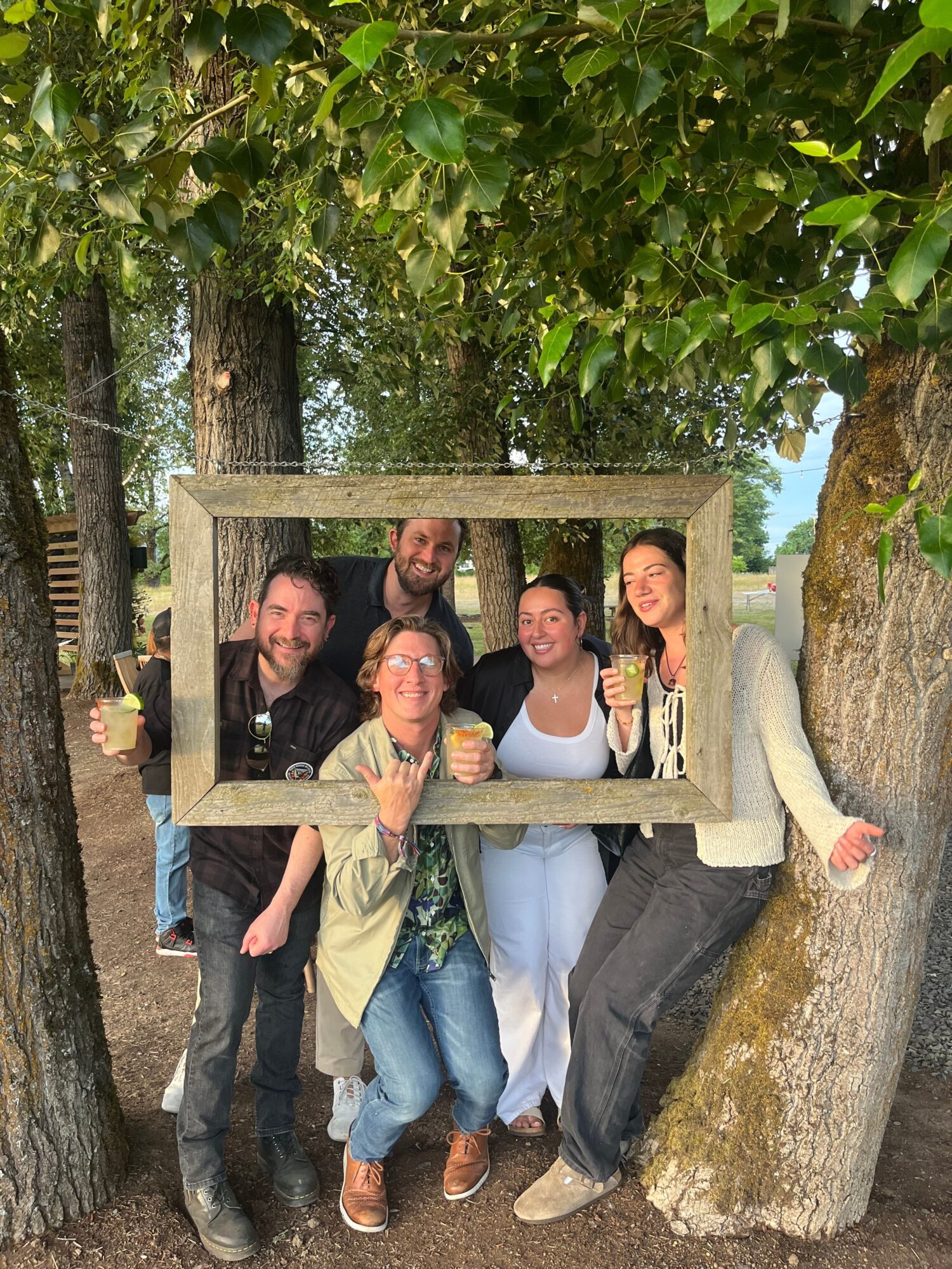 Group of five people posing outdoors, some leaning on a wooden frame, surrounded by trees and rocks.
