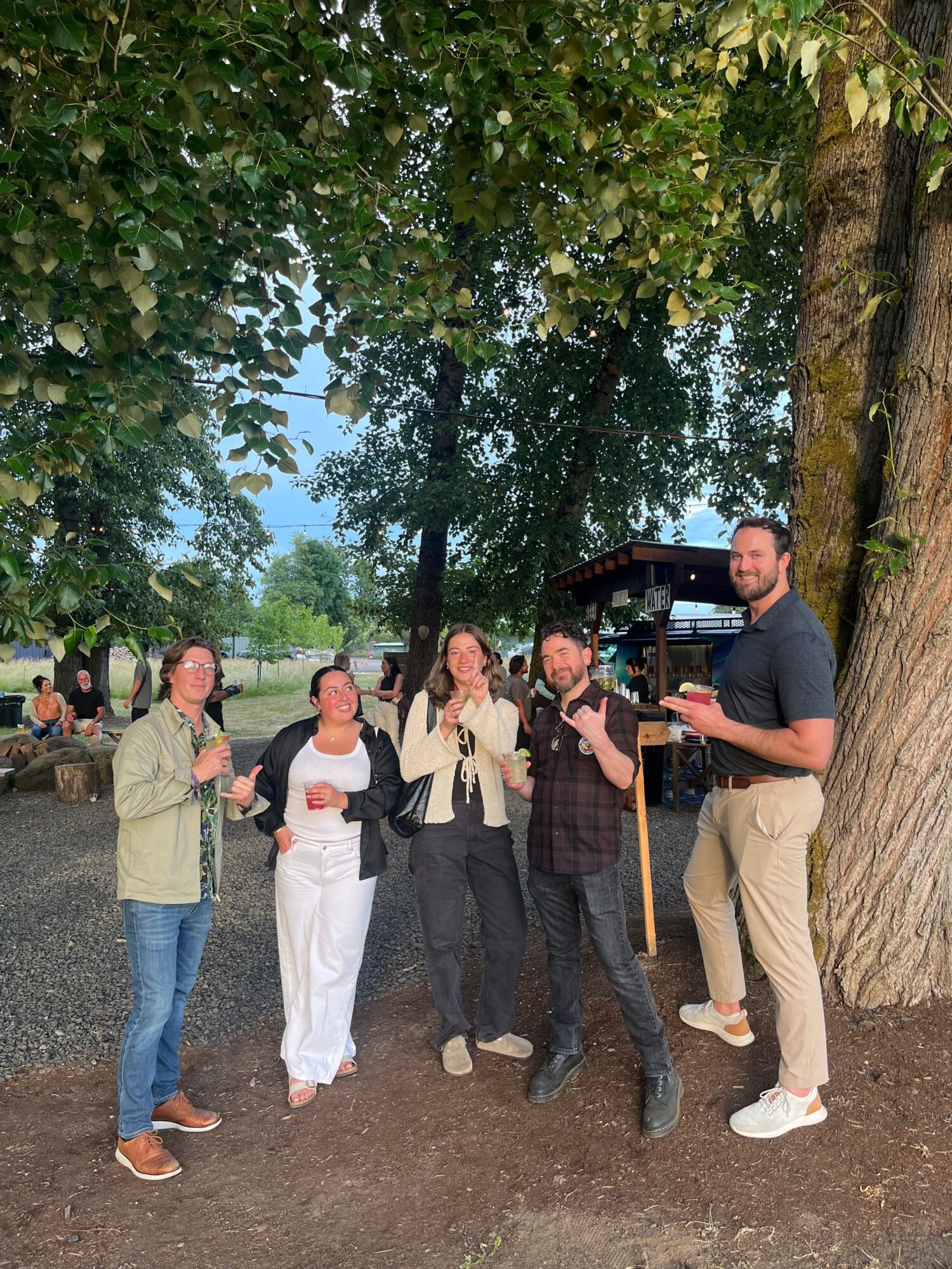 Group of six people standing outdoors near a tree, some holding drinks, with greenery and a blue sky in the background.