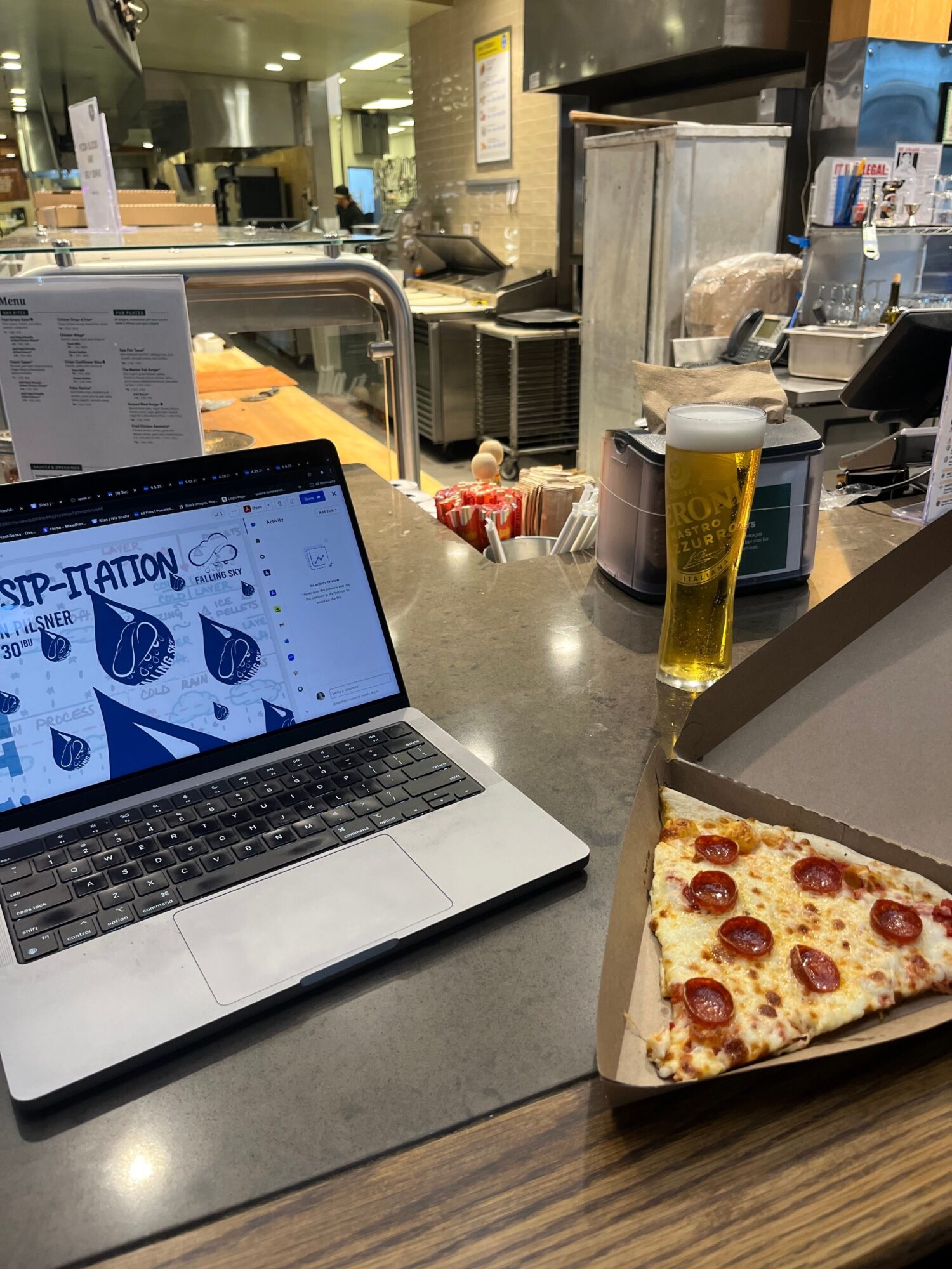 Pizza slice on a box, laptop, oil bottle, and kitchen counter with various items.