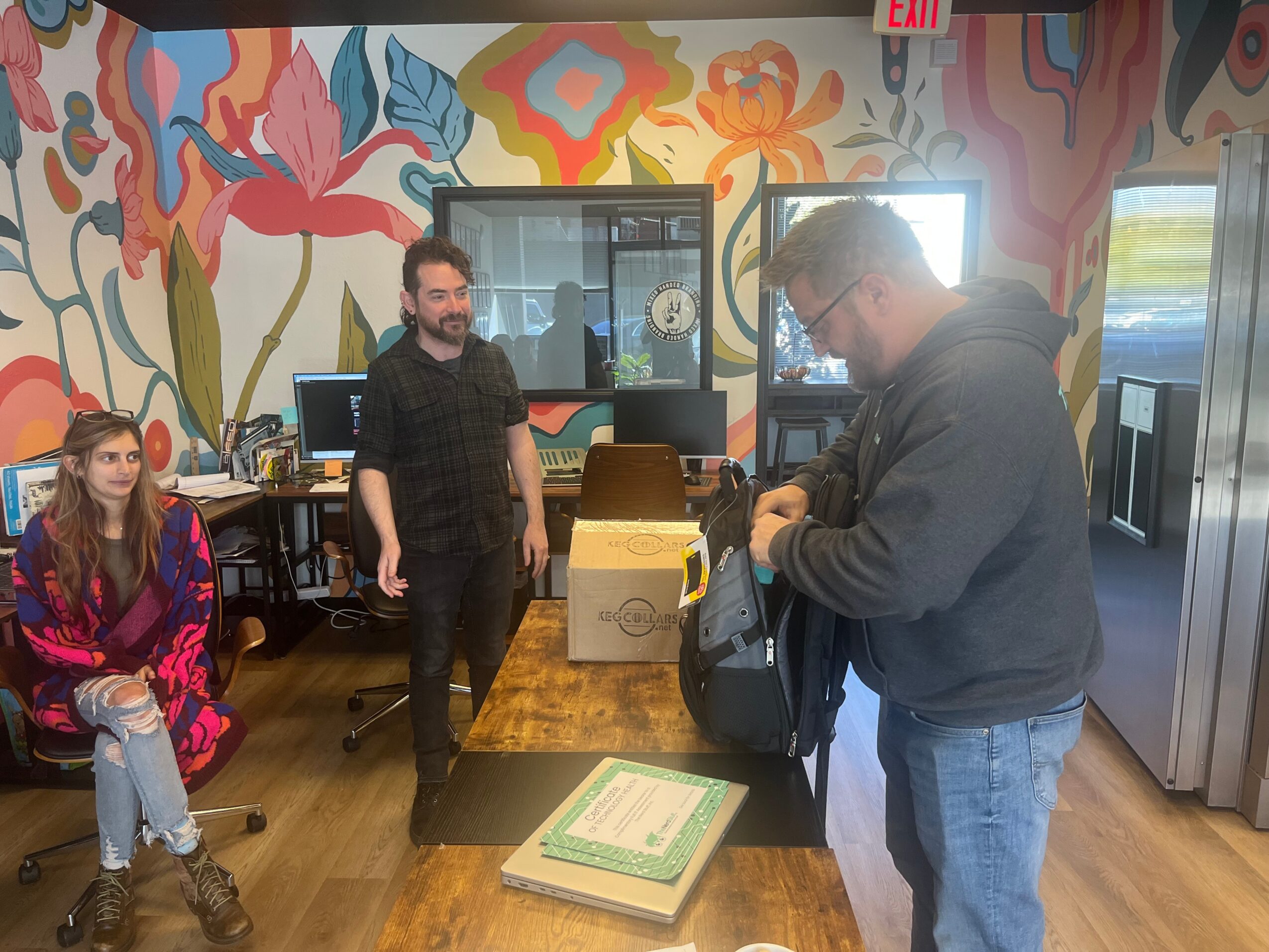 Two men and a woman in a colorful room with a mural, one man is standing, the other is opening a bag, woman sitting.