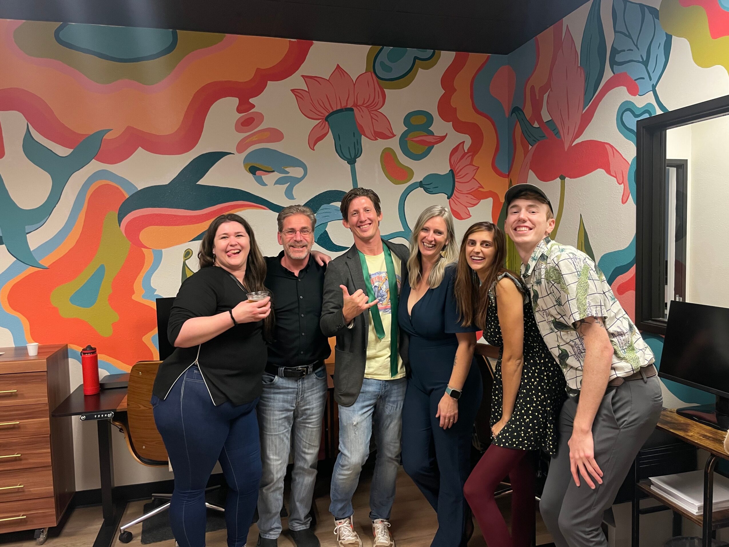 Six people standing together in a colorful room, smiling, with a vibrant abstract mural on the wall behind them.
