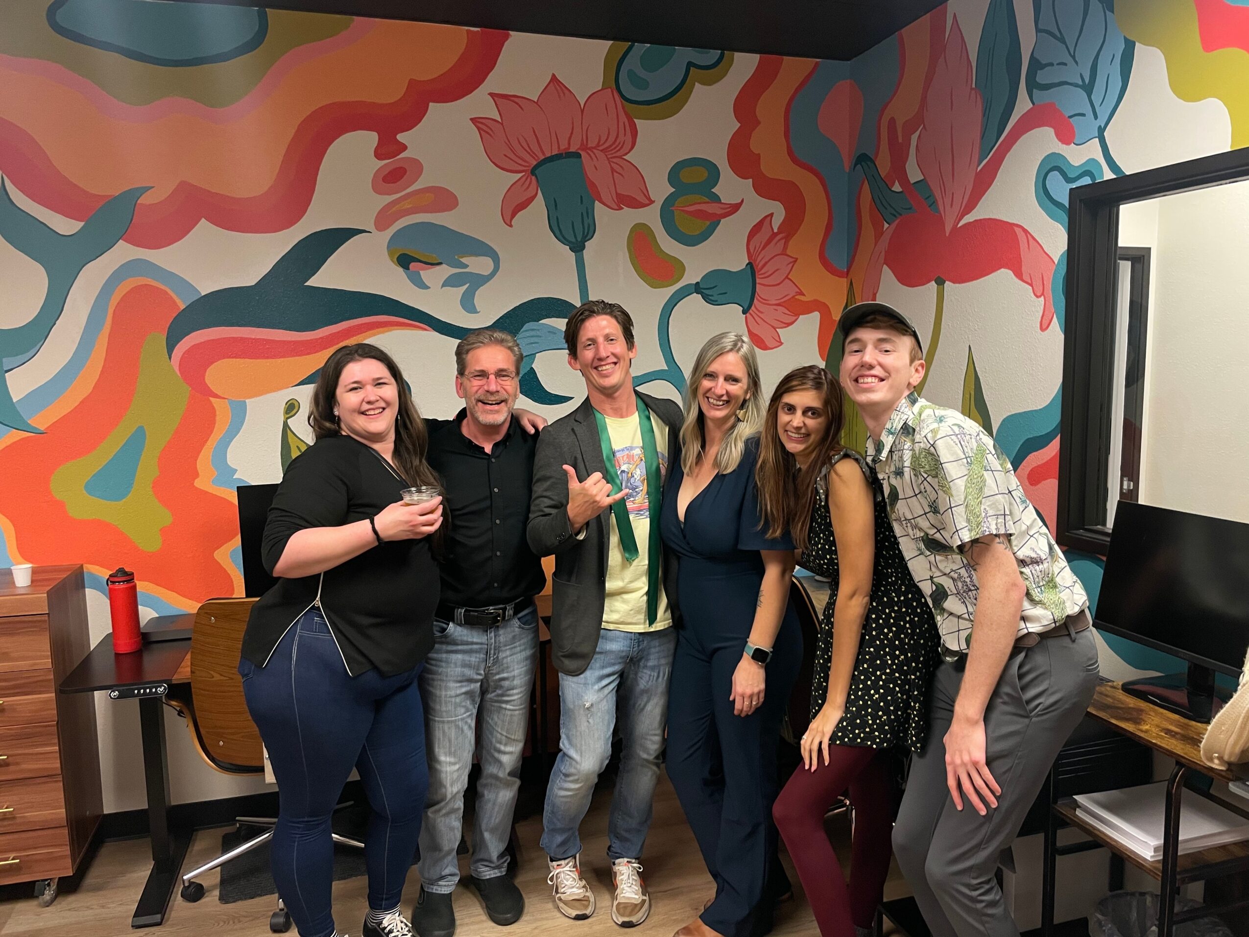 Group of six people smiling and posing in front of a colorful abstract mural, some holding drinks, in a room with a computer and guitar.