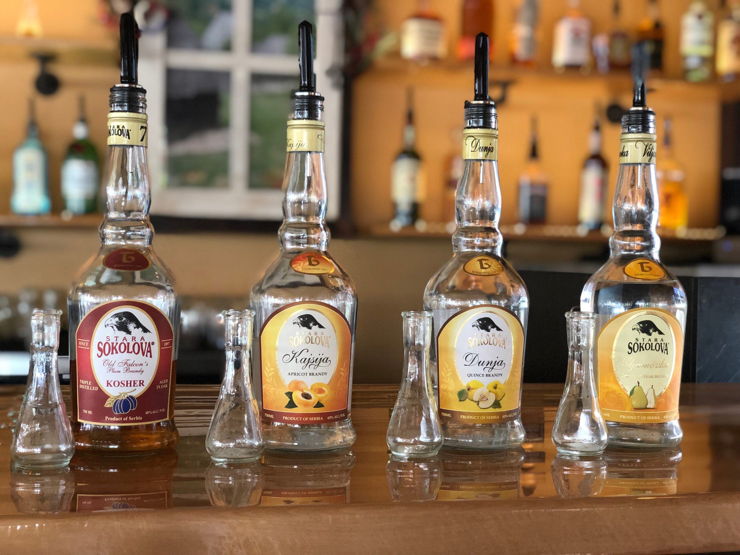 Four bottles of flavored syrup with black pour spouts on a wooden bar counter, blurred background of bottles and a window.