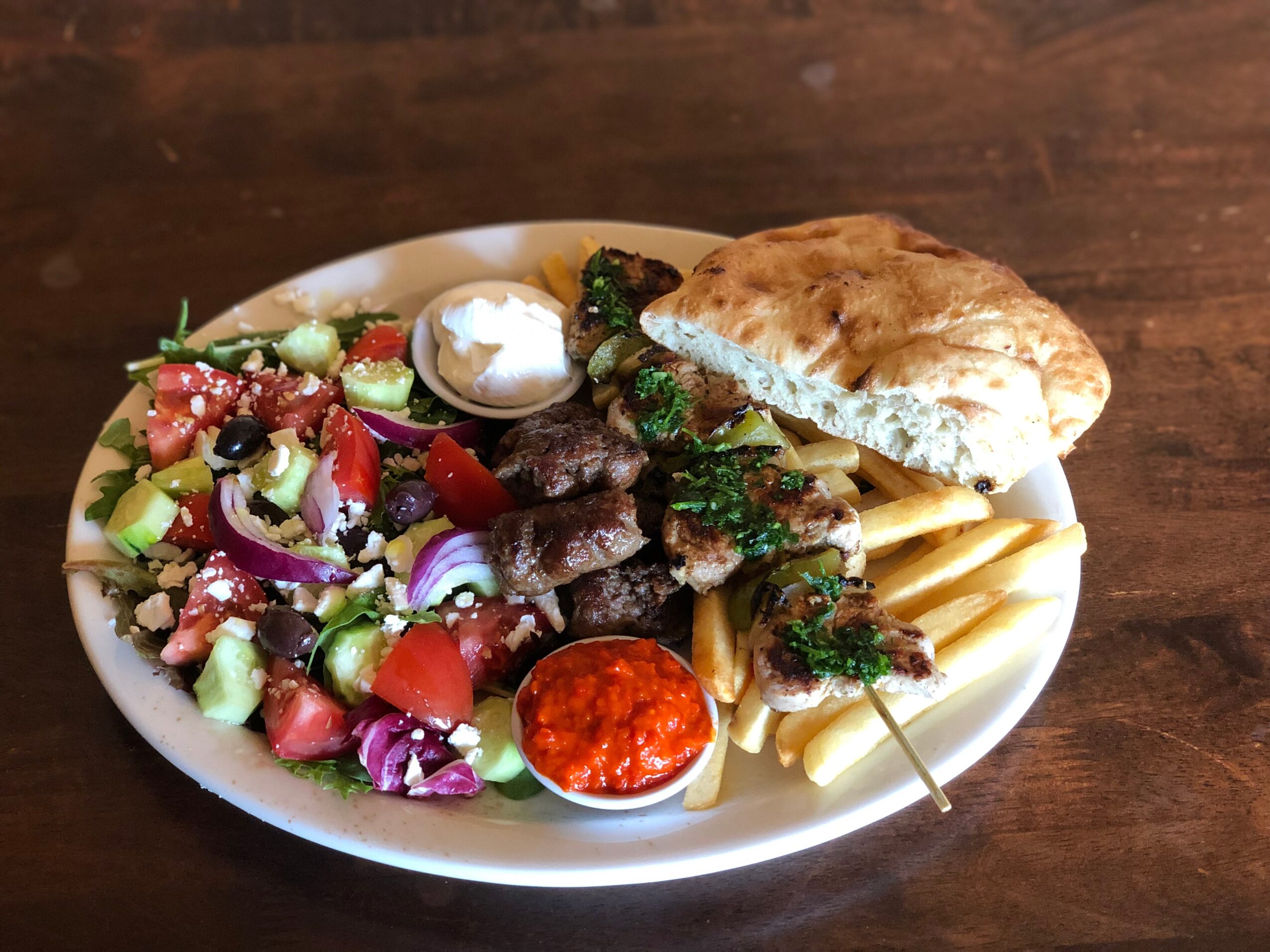 Plate with salad, grilled meat skewers, flatbread, and a small bowl of red sauce on a wooden table.