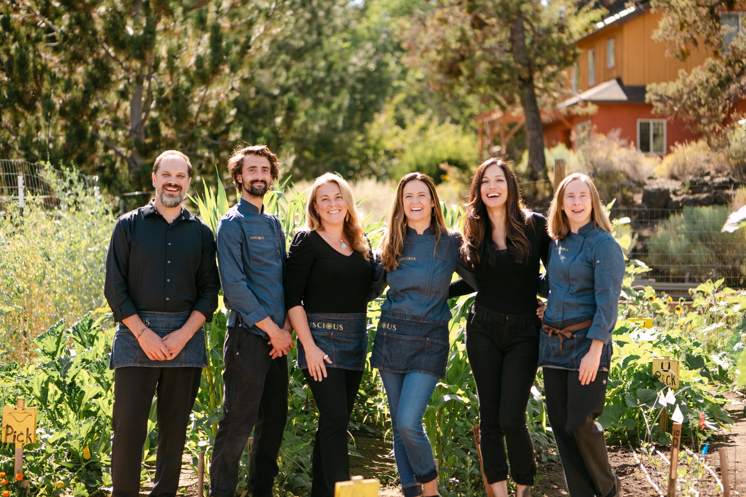 Six people standing outdoors in a garden, smiling, with trees and a house in the background.