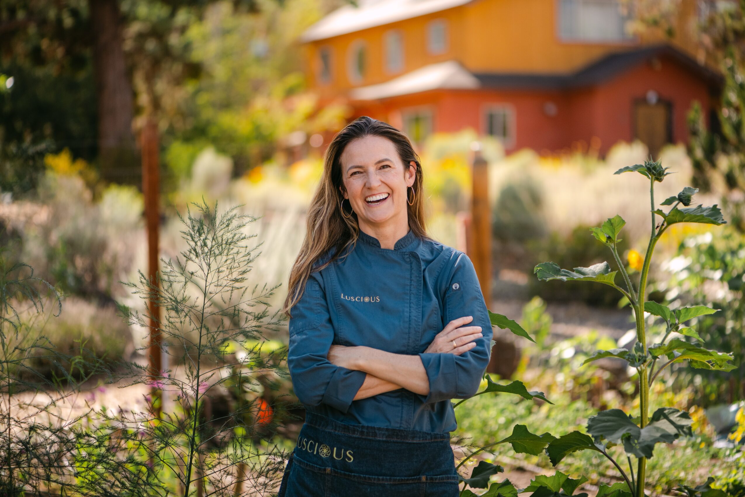 Woman smiling outdoors with arms crossed, surrounded by plants and a colorful building in the background.