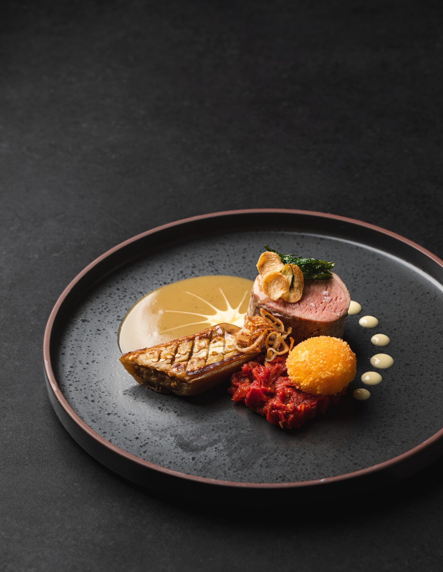 Assorted plated food with lemon slice, meat, vegetables, and sauce on a black plate, on a dark surface.