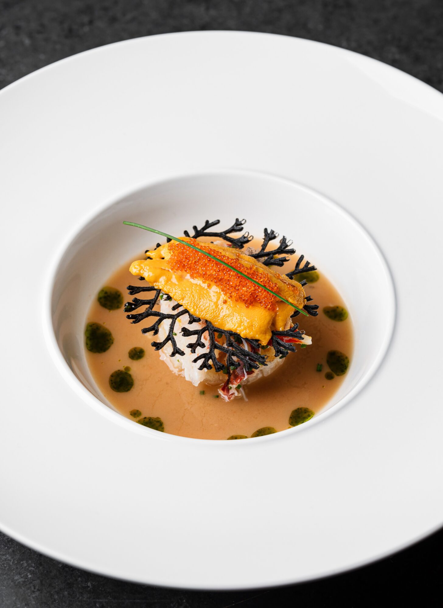 A plated dish with a piece of fried fish, black garnish, green sauce dots, and a creamy base, on a white plate.
