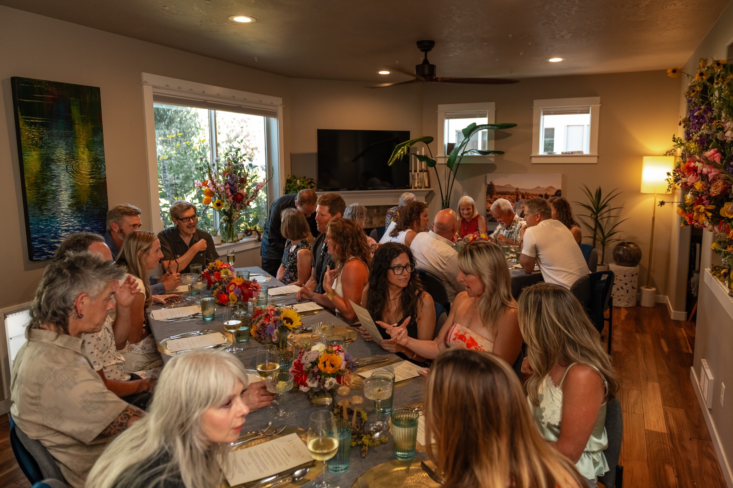 People gathered around a long dining table with flowers and food in a well-lit room, some talking and others looking at their phones.