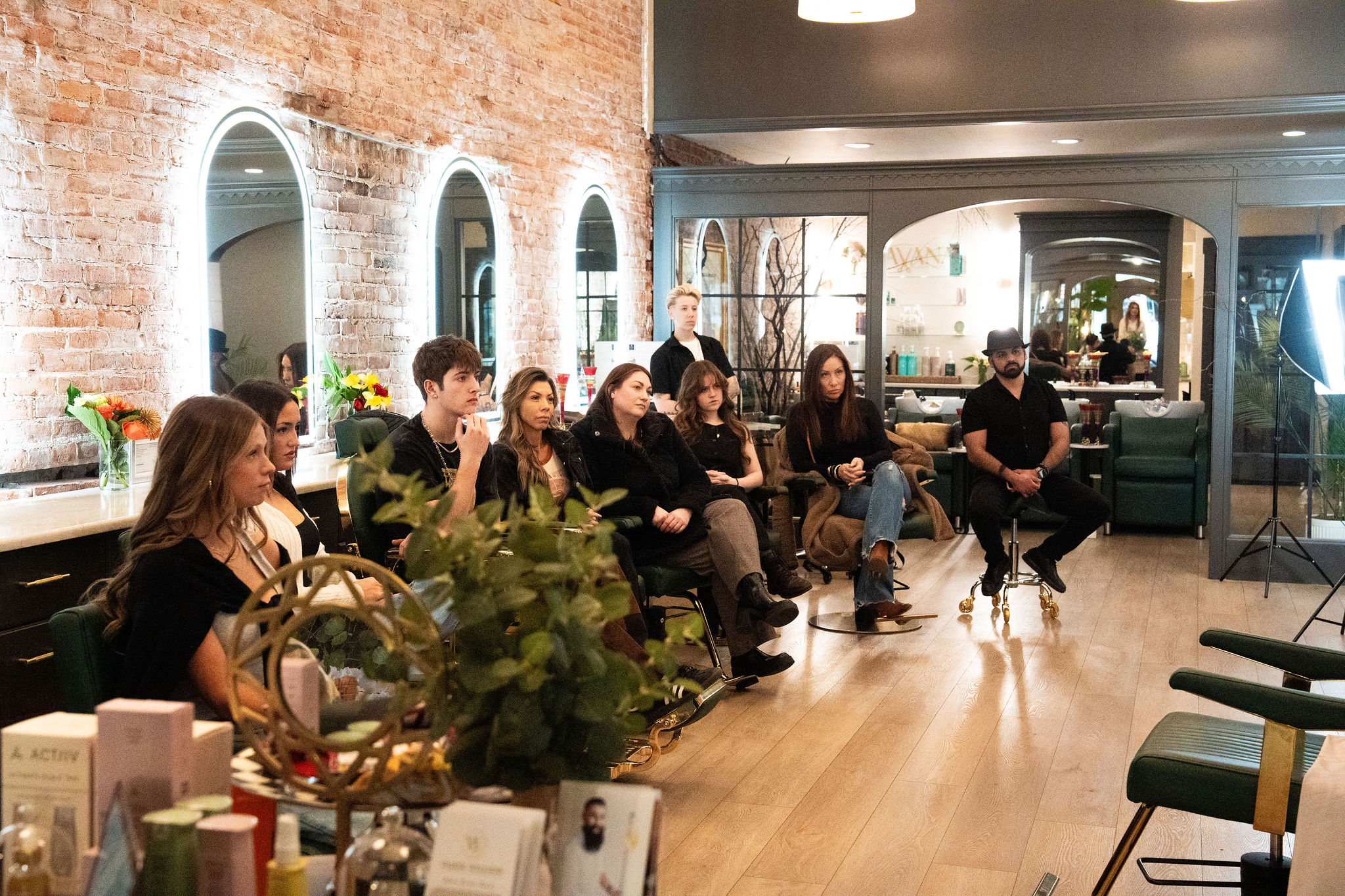 Group of people seated in a modern room with brick wall, mirrors, and large windows, participating in a discussion or presentation.
