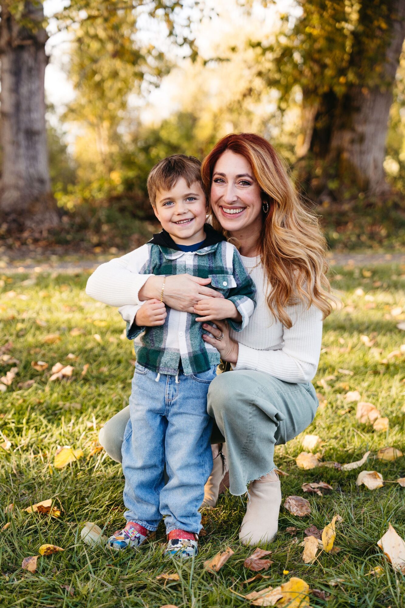 Woman and young boy smiling outdoors in a park with trees and fallen leaves, woman hugging boy, autumn setting