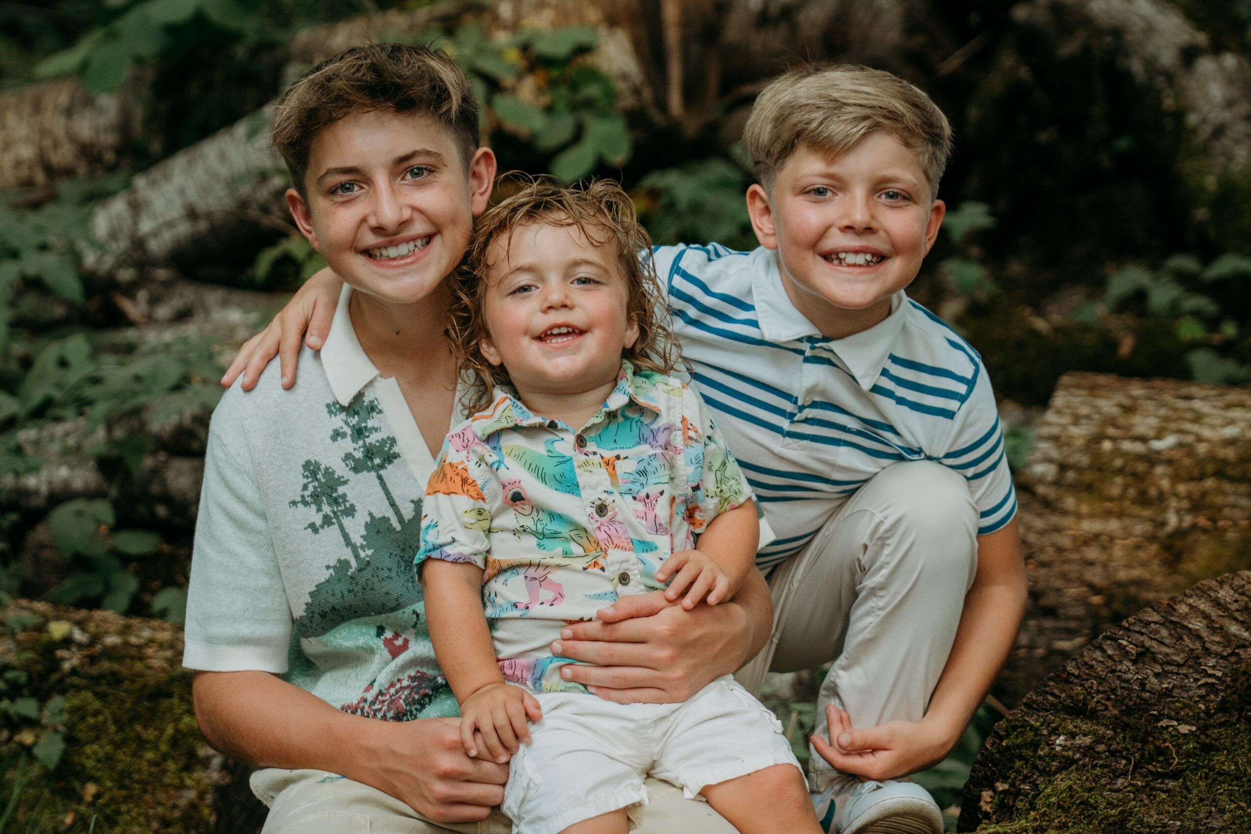 Three children smiling outdoors, sitting on logs with trees in the background.