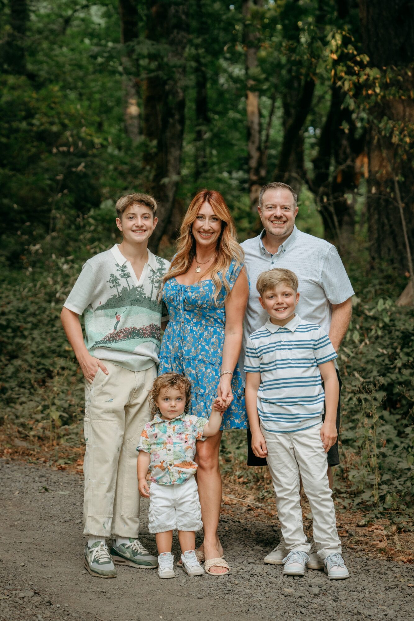 Five people standing outdoors in a forest, smiling, with trees and greenery in the background.