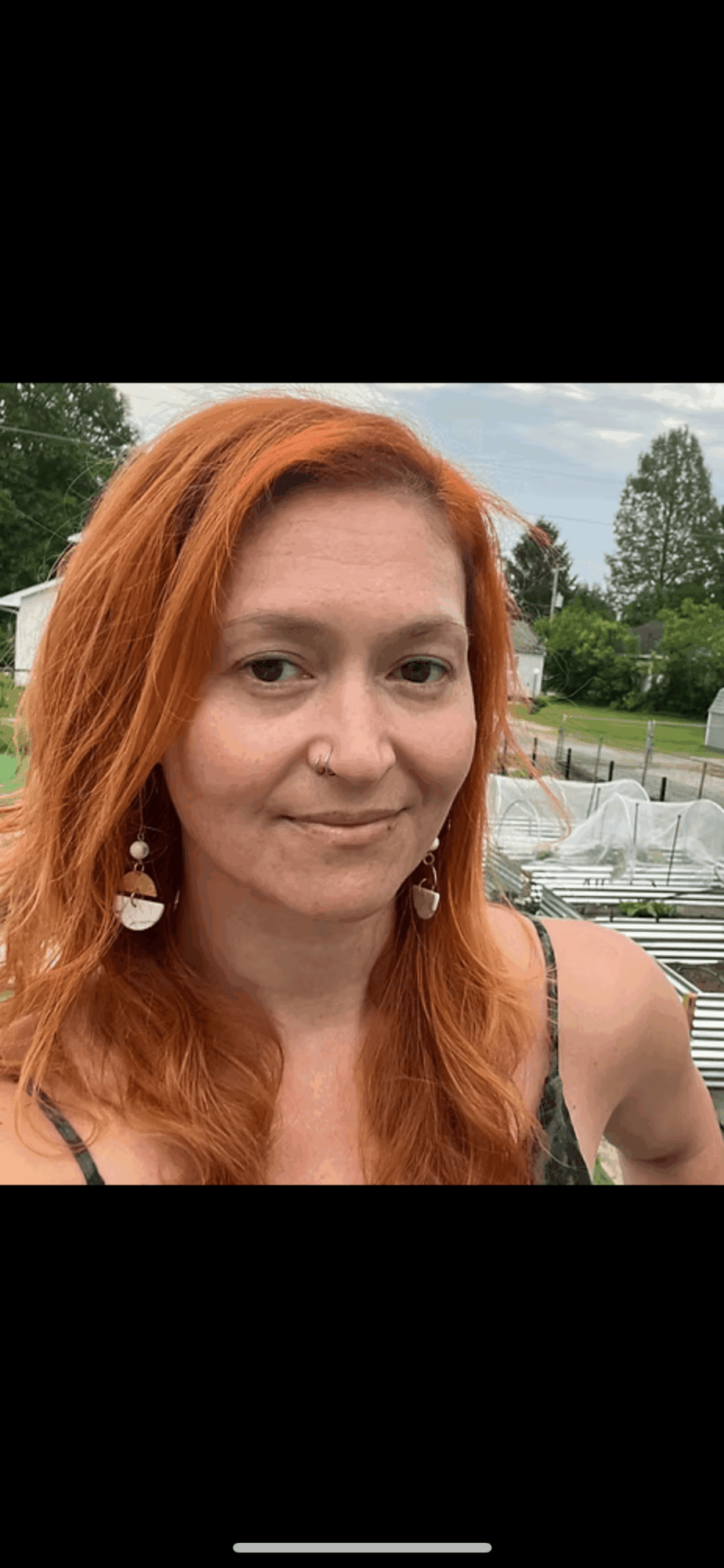 Woman with red hair outdoors, trees and a cloudy sky in background, smiling slightly, wearing earrings and a sleeveless top.