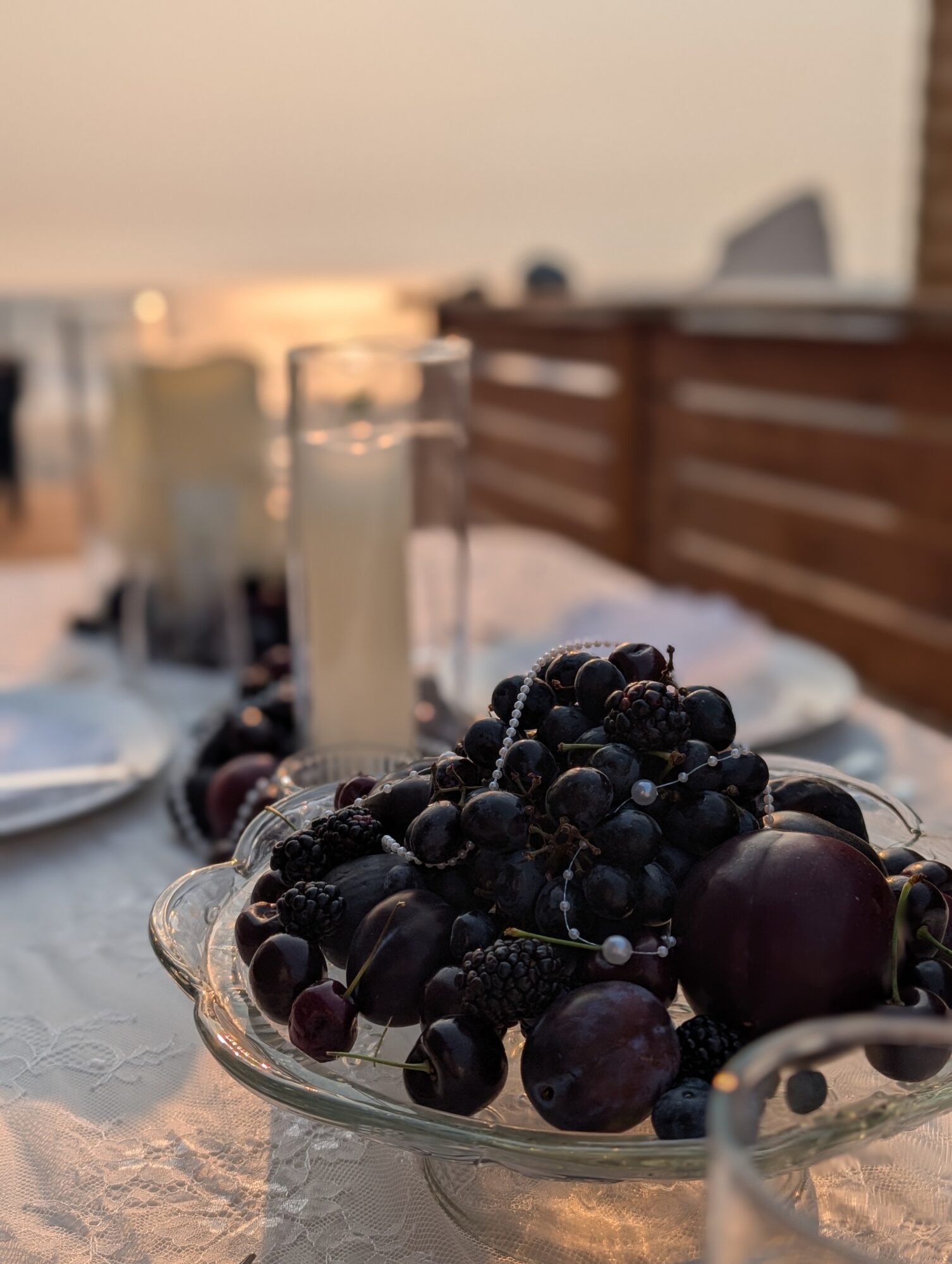 A glass dish filled with dark grapes on a table set for a meal, with candles and glasses in the background.
