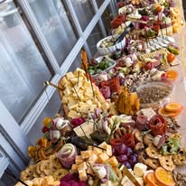 A long table with various cheeses, fruits, and snacks arranged for a buffet or party, seen from the side.
