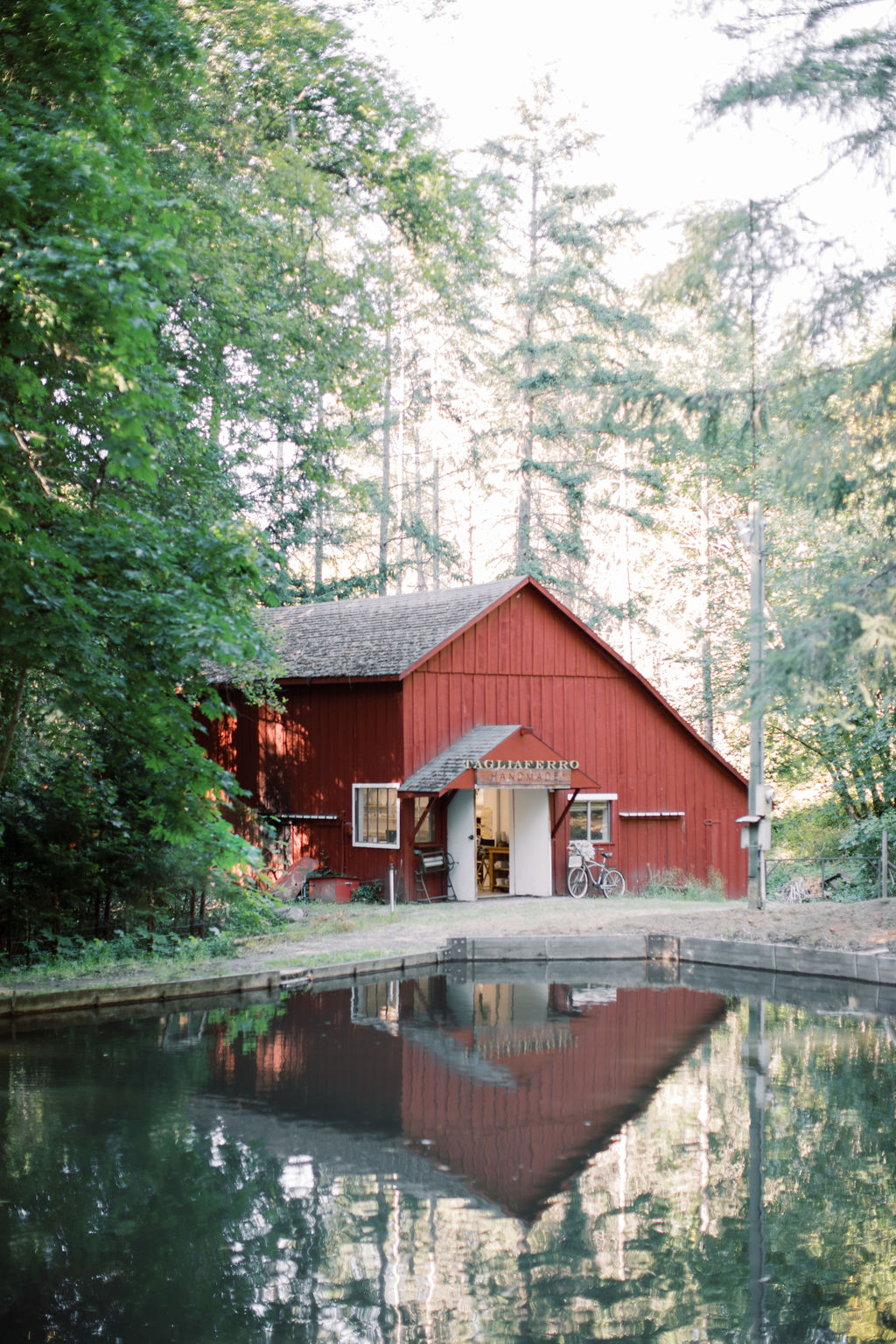 Red barn with a gray roof beside a pond, surrounded by trees, reflecting in the water, daytime.