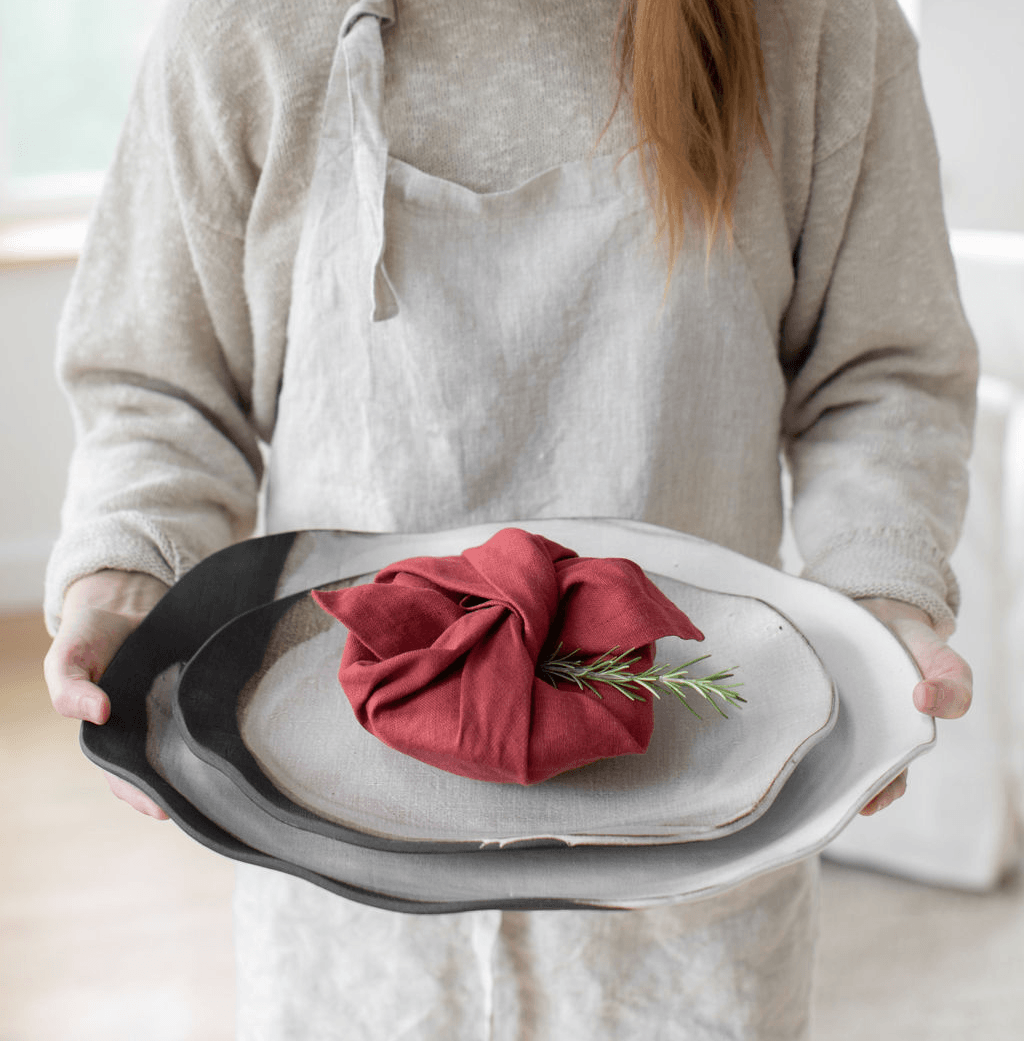 Person holding a tray with a red cloth-wrapped item and a sprig of greenery, wearing a beige apron and sweater.