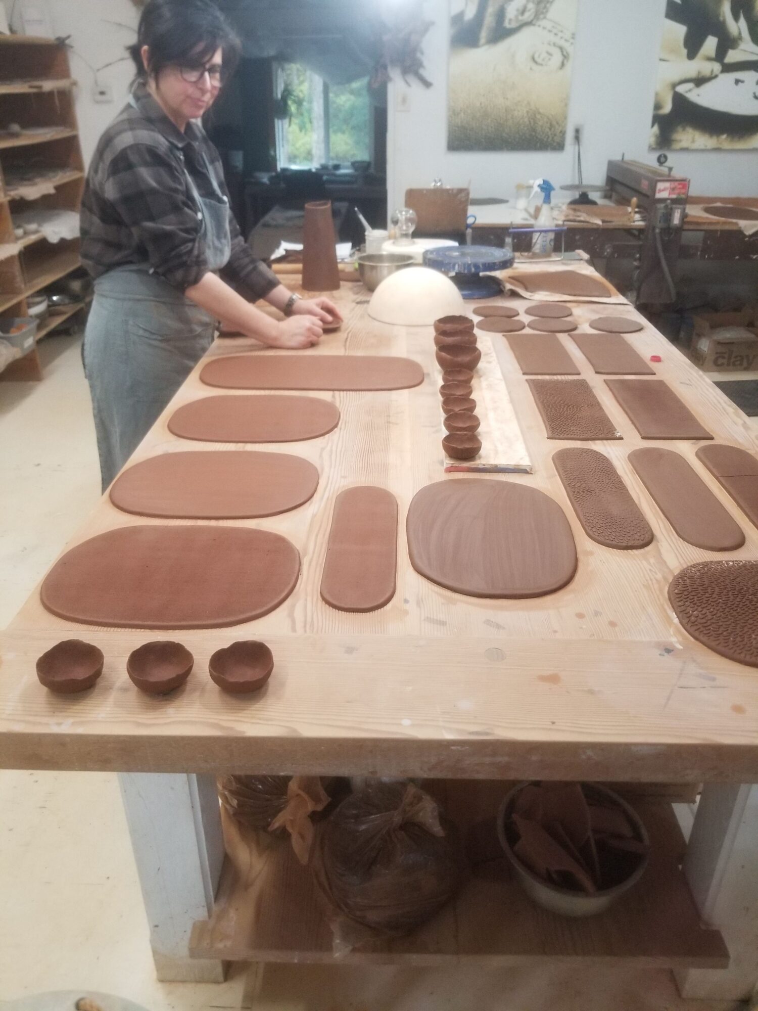 Person working on a long wooden table with various oval and circular clay or dough pieces, in a workshop setting.