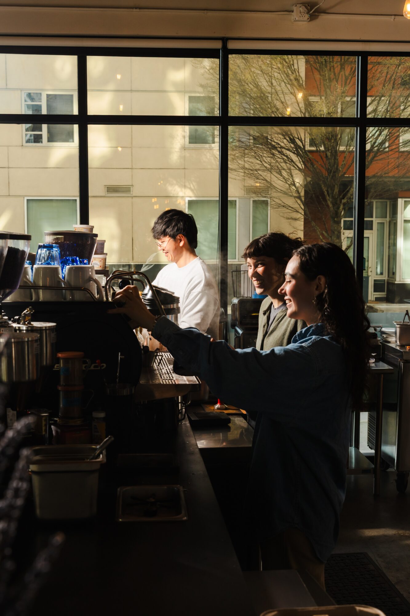 People working behind a counter in a cafe with large windows and natural light, trees visible outside.