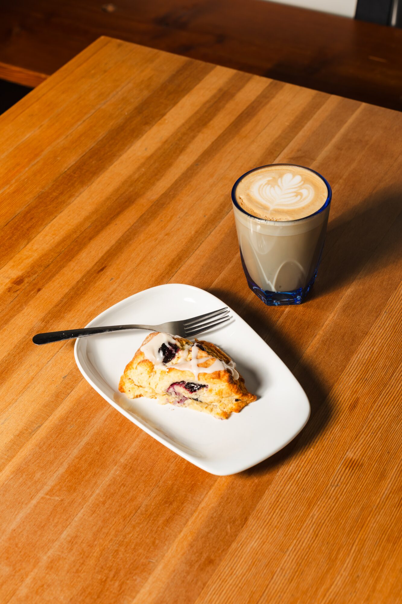 A partially eaten slice of berry-topped dessert on a white plate with a fork, and a latte in a glass on a wooden table.