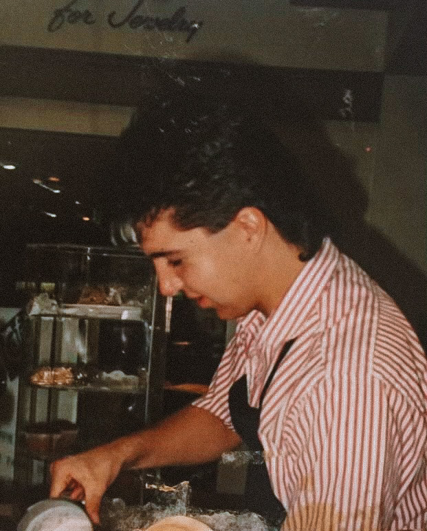 Young man with dark hair in a striped shirt, looking down, in a dimly lit setting with food display in background.