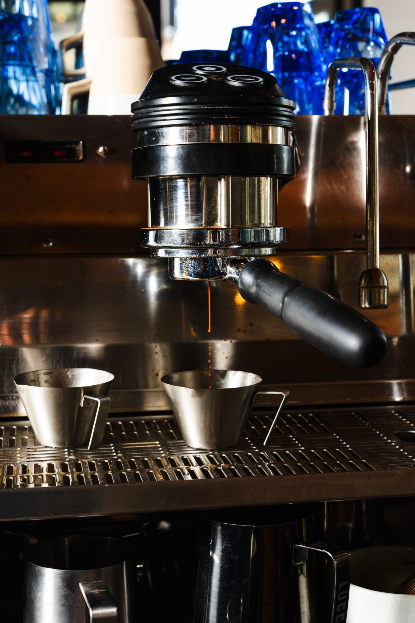 Espresso machine with portafilter and two metal cups on drip tray, blue bottles in background.