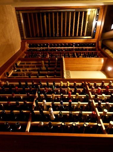 Wine cellar with wooden racks filled with bottles, a wine fridge, and warm lighting, viewed from the entrance.