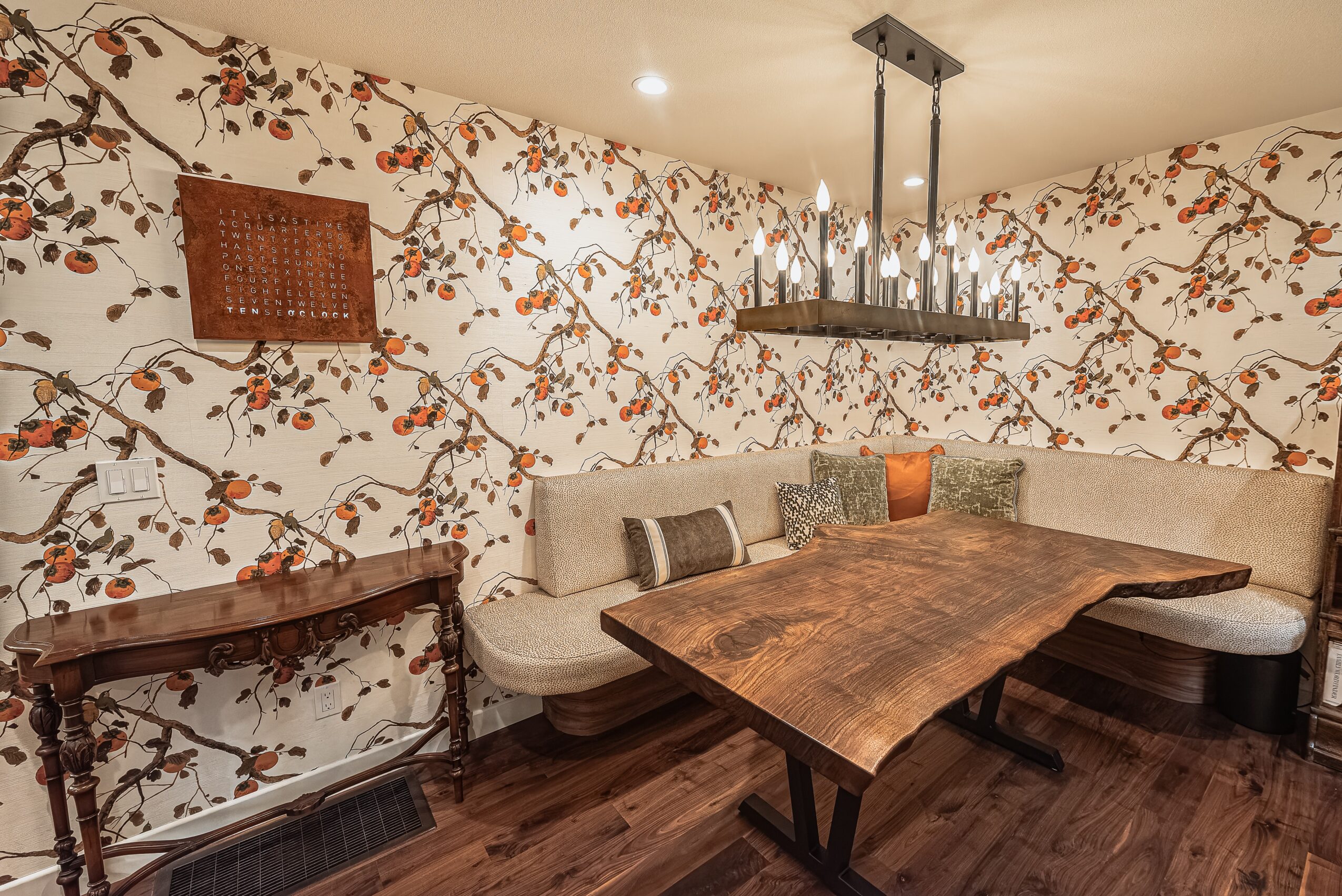 Corner of a room with floral wallpaper, a curved sofa with cushions, a wooden table, and a chandelier.