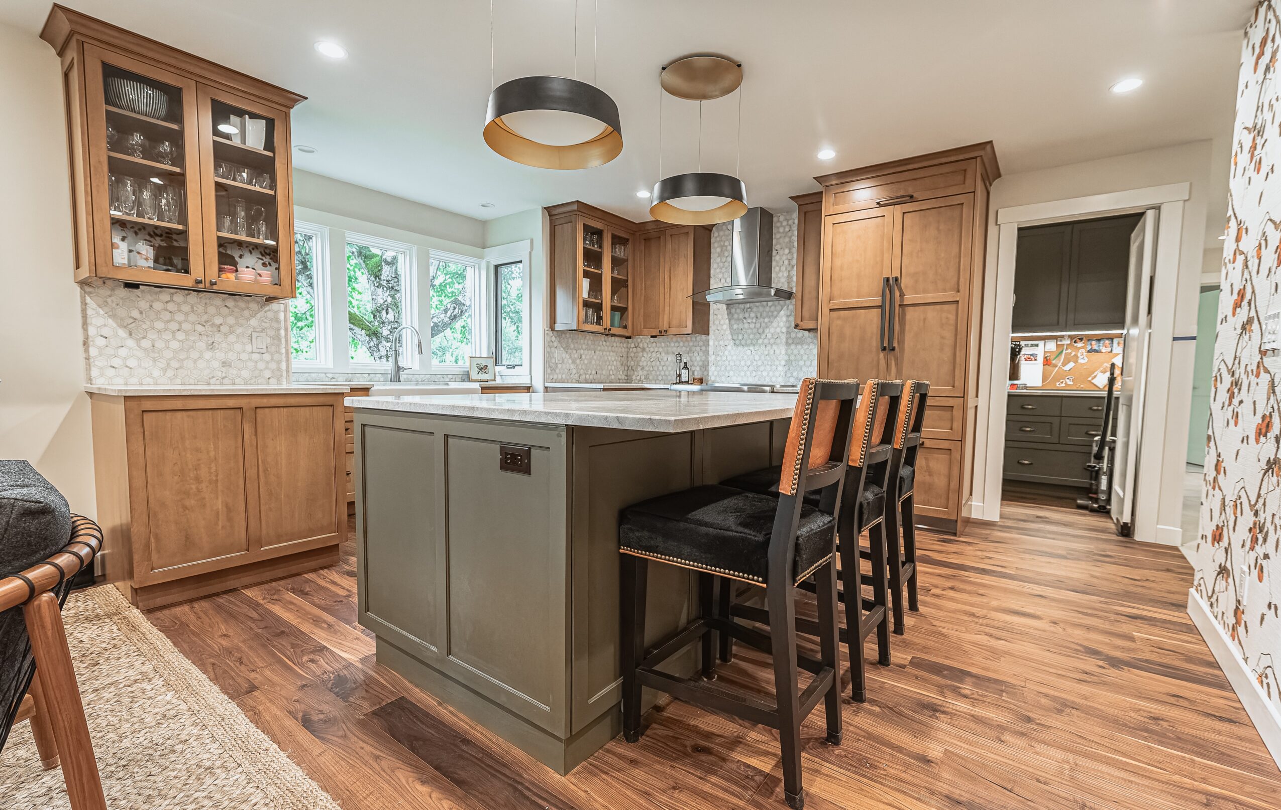 Kitchen with wooden cabinets, an island with barstools, and hardwood flooring, illuminated by ceiling lights and natural light from windows.