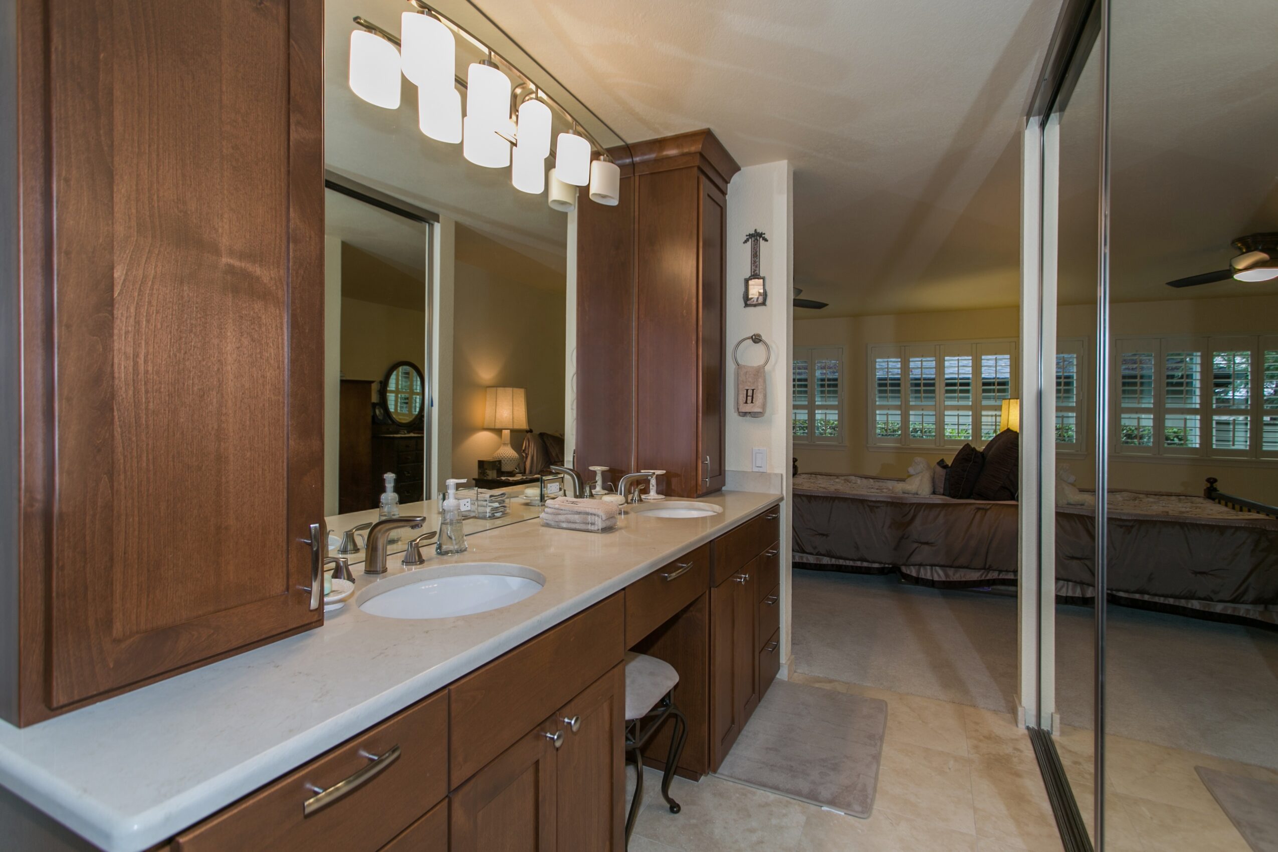 Bathroom vanity with double sinks, wooden cabinets, large mirror, and modern light fixture, leading to a living room with couches and windows.