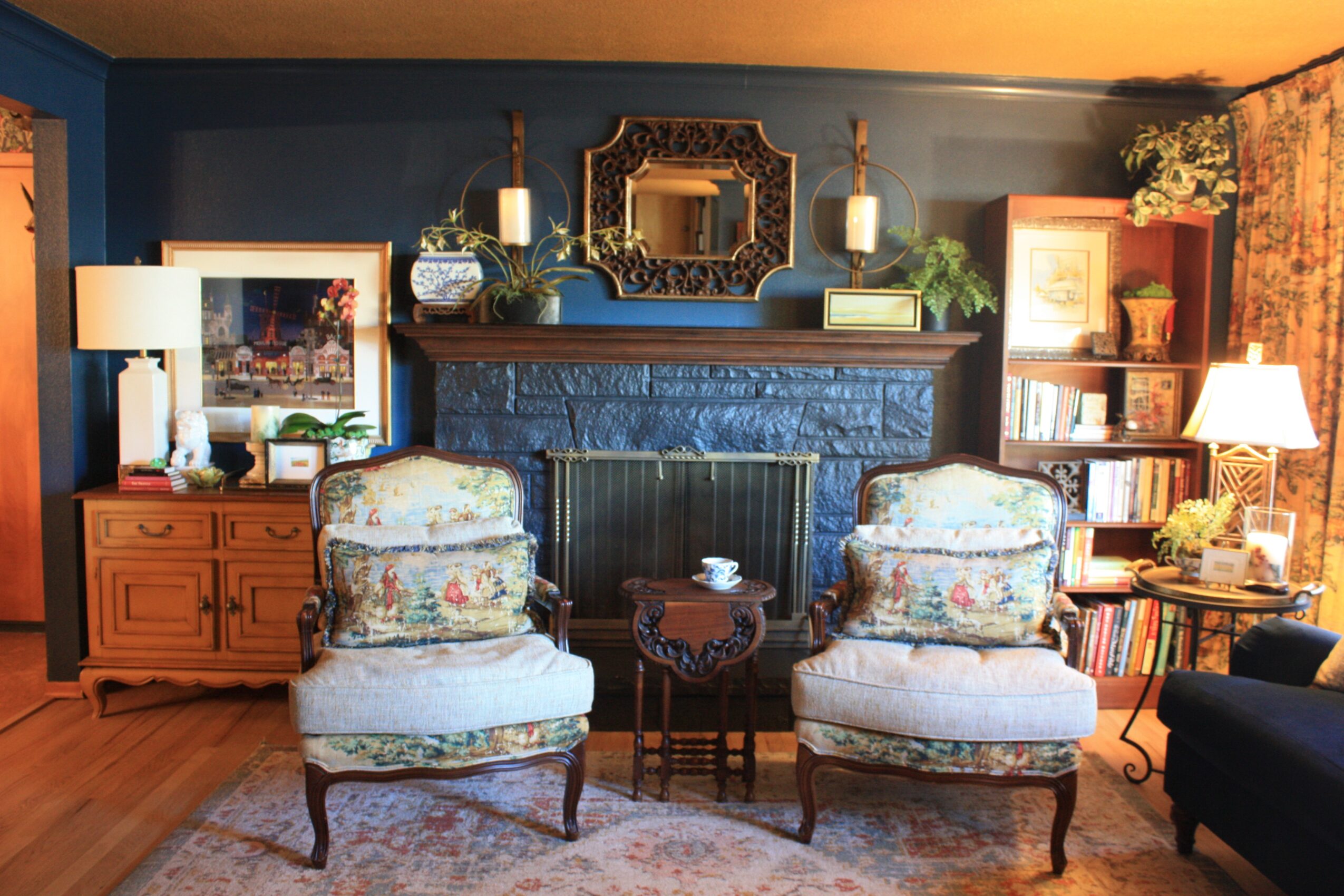 Living room with blue wall, fireplace, two floral armchairs, wooden side tables, and various lamps and decorations.